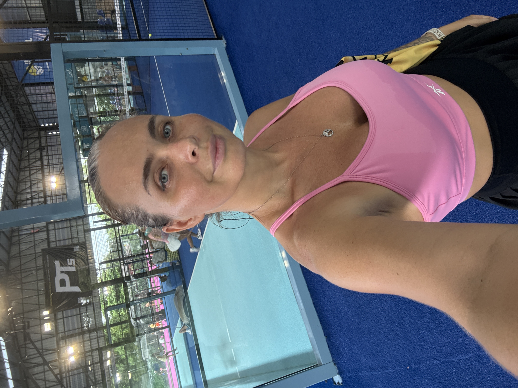 The image shows a woman taking a selfie indoors, likely at a padel tennis club in Ban Bang Rak, Thailand. She is wearing a pink athletic top and a black skirt. In the background, several people are playing padel tennis on blue courts, enclosed by glass walls. There are also banners and sports equipment visible. The lighting appears to be artificial, suggesting an indoor facility.