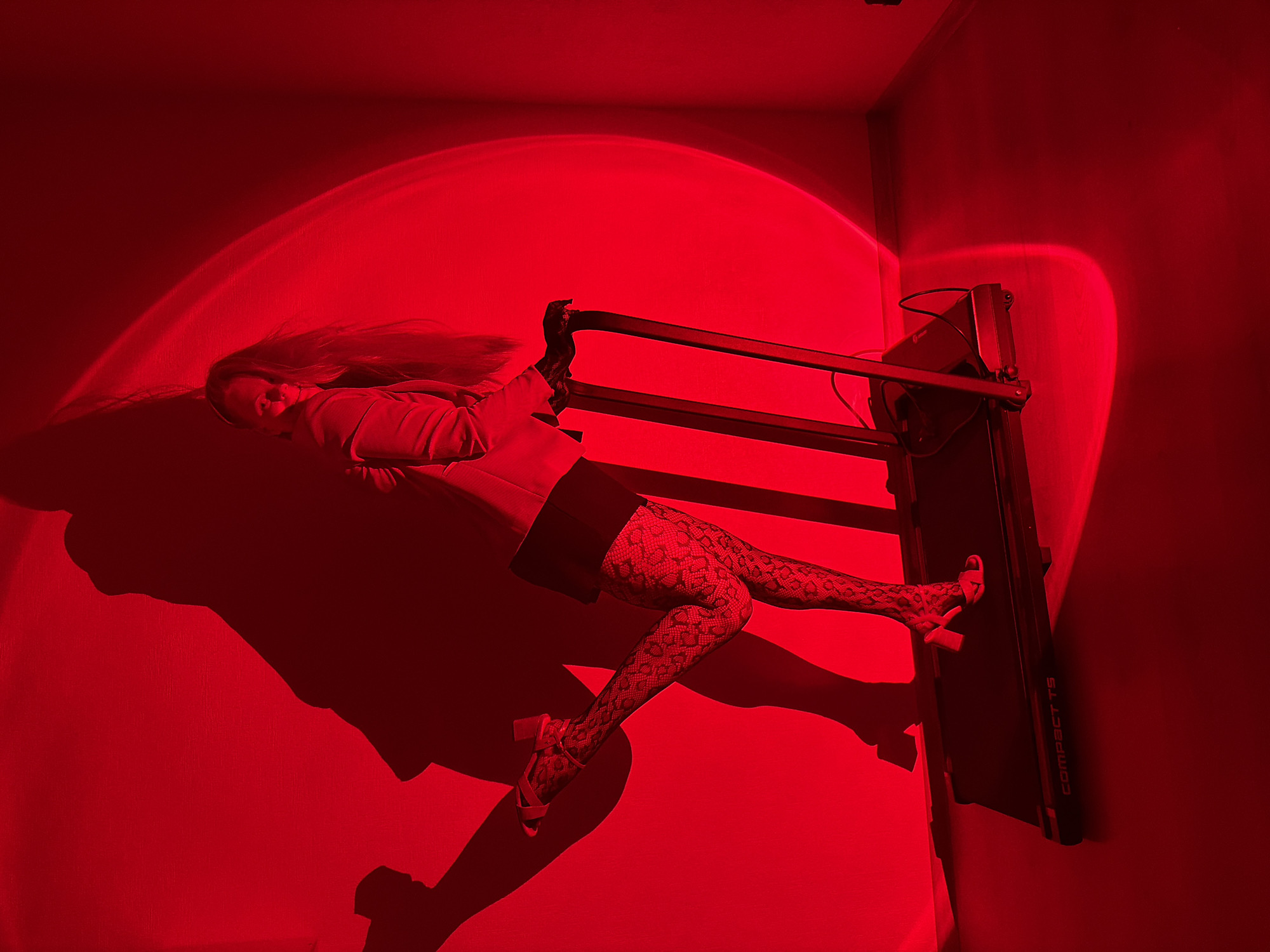 A woman is posing on a treadmill in a room illuminated by red light. Her body is angled towards the viewer, with one leg extended backward and resting on the treadmill. She is wearing a blazer, a short skirt, patterned stockings, and high-heeled sandals. The lighting casts a dramatic shadow of her on the wall behind her, creating an abstract silhouette. The room appears to be a simple interior space with plain walls and floor. The image does not contain any information that would allow for the identification of a specific location in Izhevsk, Russia.