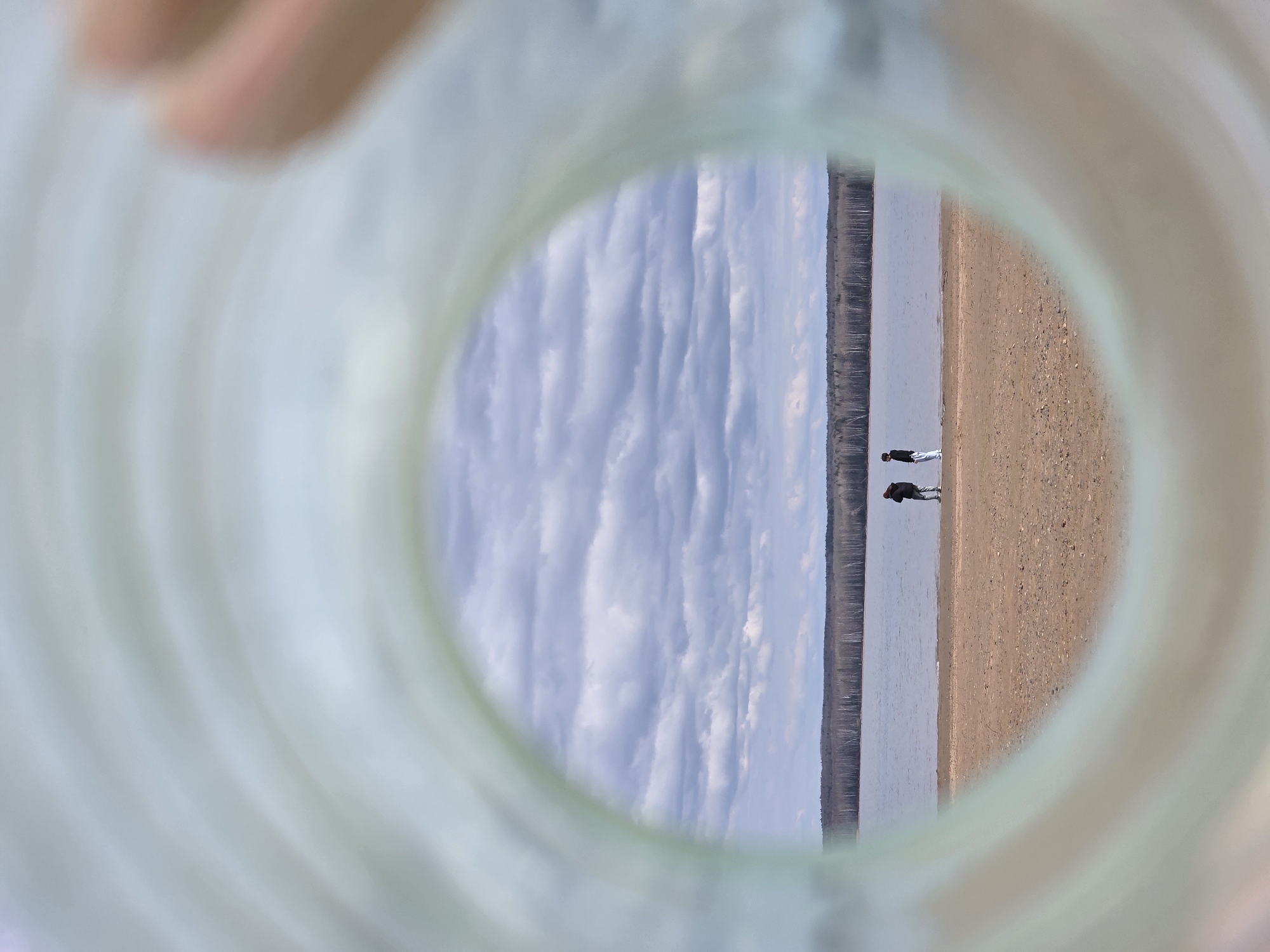 This image presents a landscape scene viewed through a blurry, translucent circular opening, resembling the neck of a bottle or tube, with a light green tint. The main scene is vertically oriented within this frame, depicting a serene natural environment.
In the background of the framed view, a sky with prominent, elongated white clouds against a pale blue backdrop dominates the upper portion. Below the sky, a distant, dark treeline (likely a forest) runs horizontally, bordering a calm body of water, possibly a wide river or lake. On the opposite side of the water, a sandy or pebbly shore extends.
Two small, dark figures, appearing to be people, are visible on this sandy shore, standing near the water's edge. Their distant placement and small size make their exact activity indistinct, but they appear to be enjoying a stroll or simply observing the surroundings.
The time of day appears to be daytime, possibly late morning or afternoon, given the bright illumination. The weather seems fair, characterized by the visible clouds. There is no visible text in the image. The location context provided is Chaikovsky, Russia, which aligns with the depiction of a wide river or lake (like the Kama River) bordered by forests and a sandy shore. The unique framing creates a sense of looking out from a confined space into a vast, open landscape.