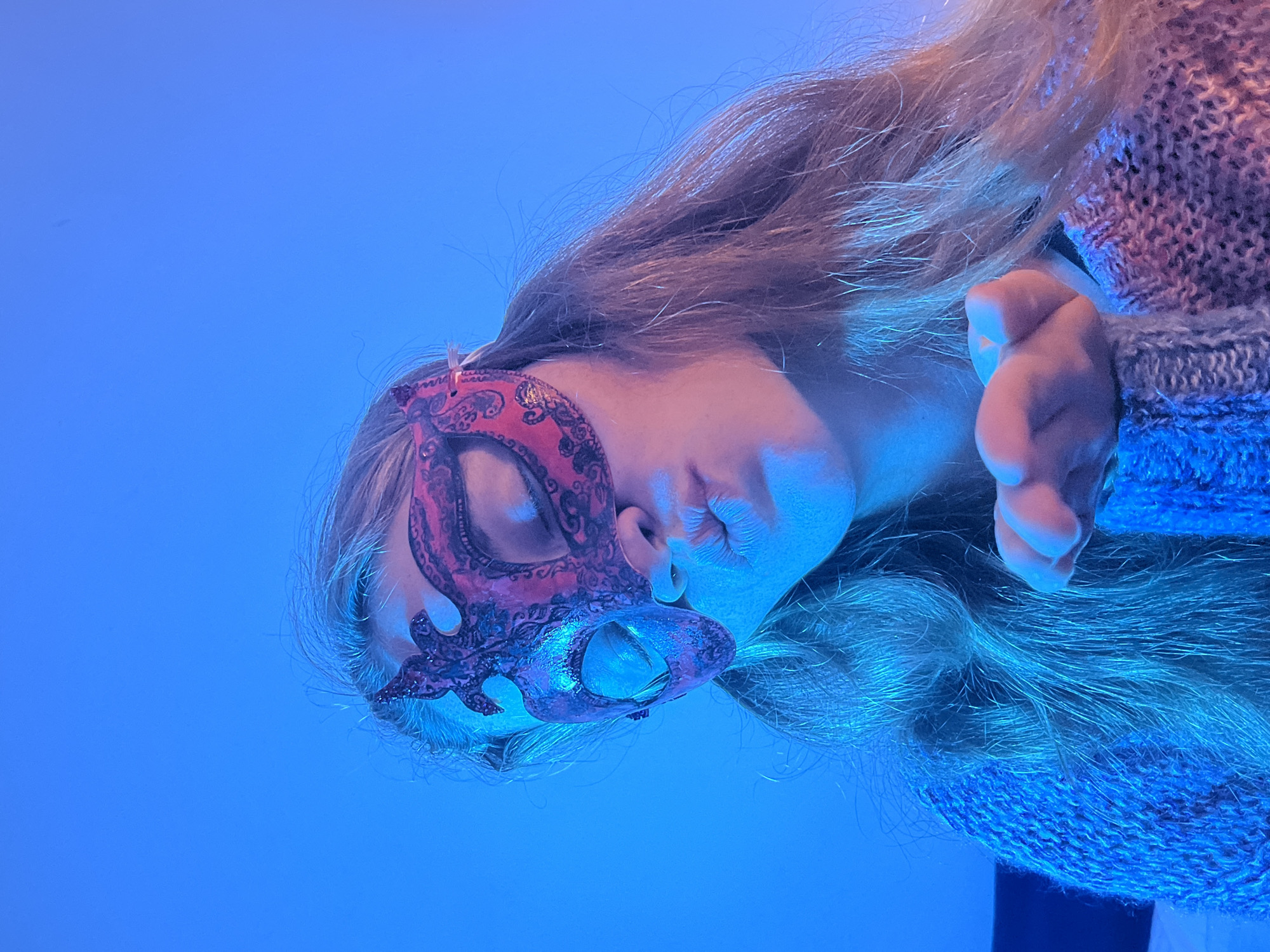 A female faces left wearing a decorative mask over her left eye, with her head tilted, possibly asleep. The lighting is predominantly blue. She wears a light-colored knitted sweater. There is no indication this photo was taken in Budva, Montenegro.