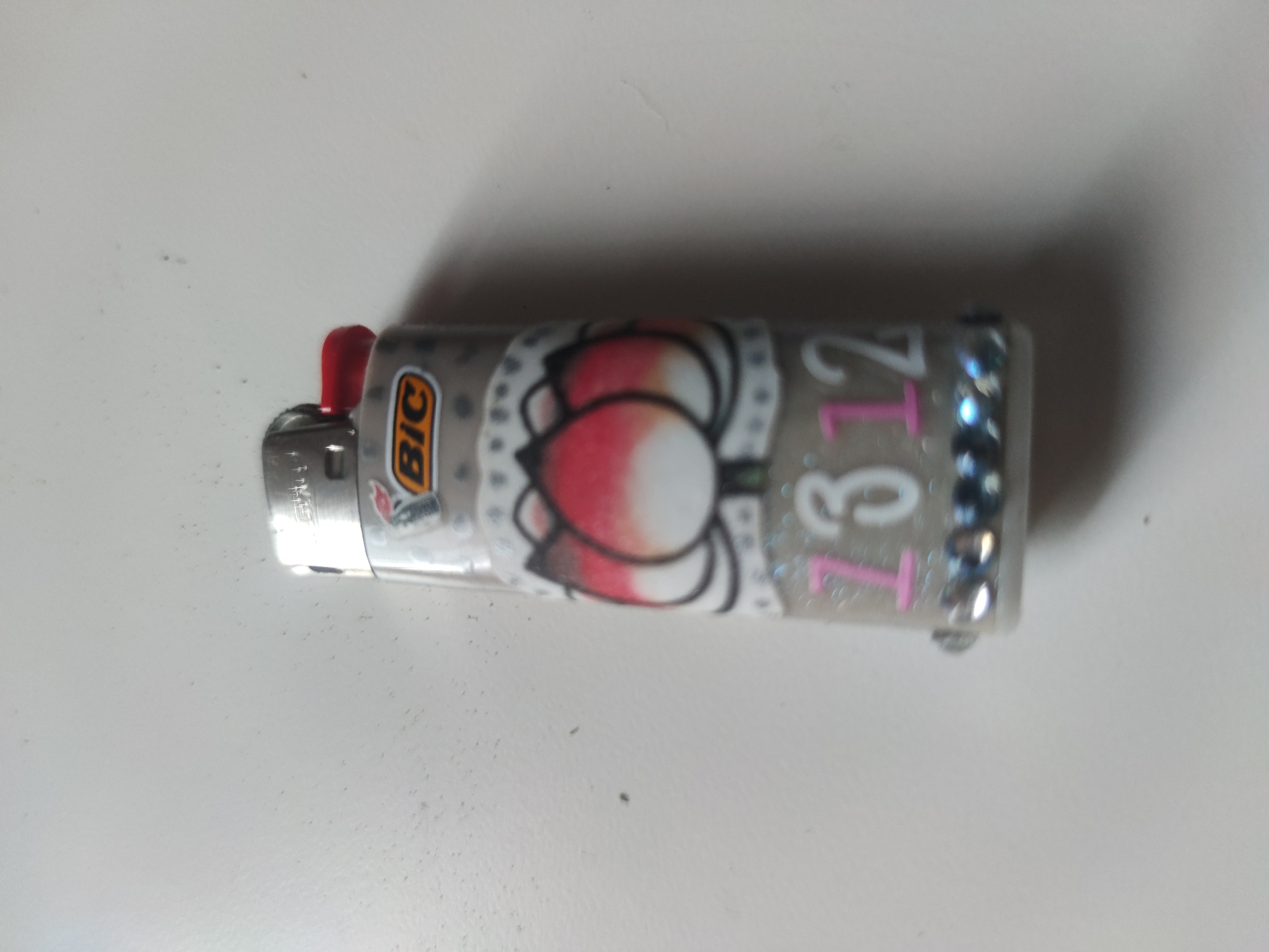 A Bic lighter sits on a white surface. The lighter is decorated with a floral design, the numbers "7312" in white with pink accents, and a row of small rhinestones near the top. The lighter's red spark lever is visible.