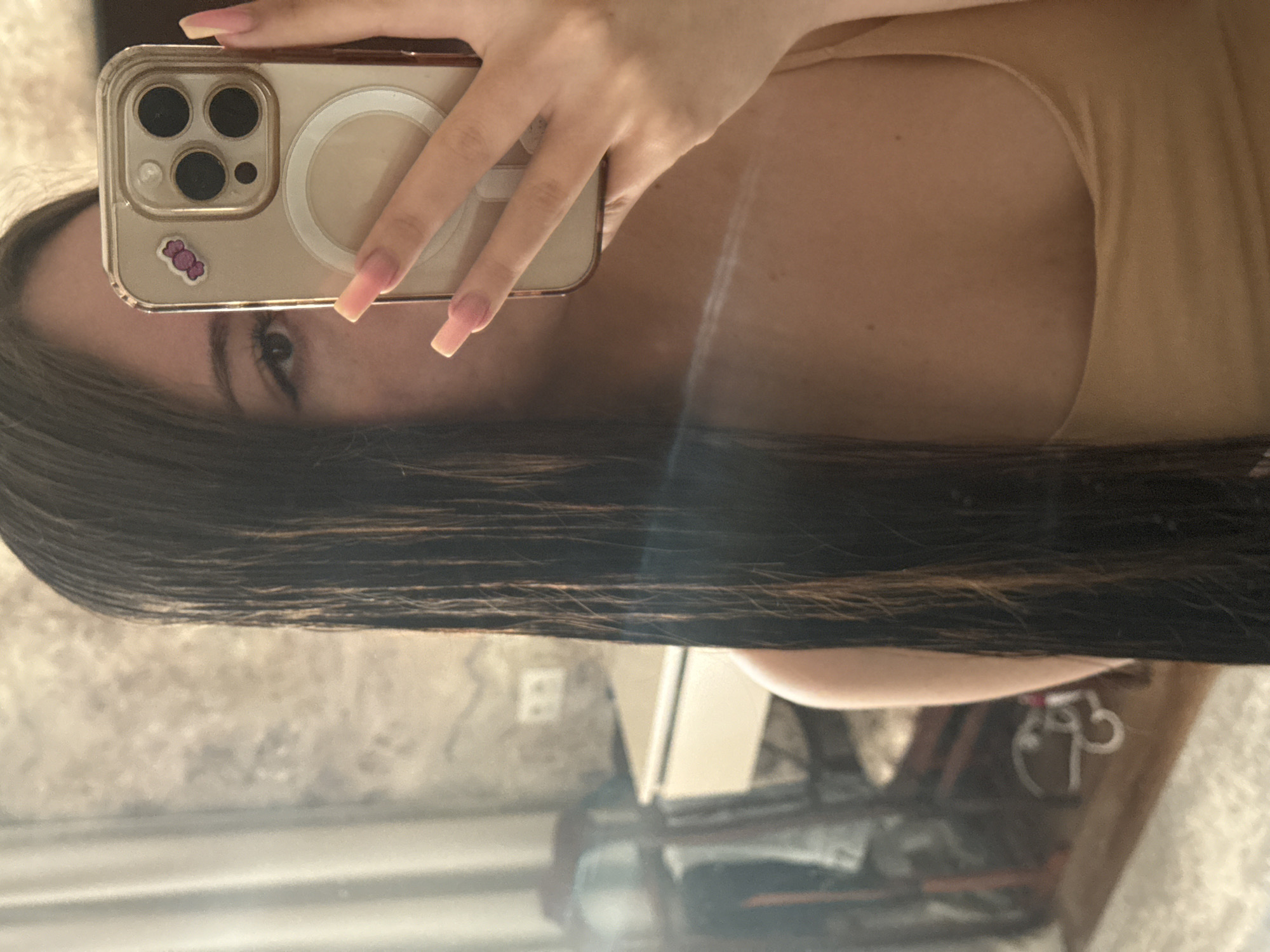 A selfie taken in an interior setting, possibly a residence, featuring a female individual holding a smartphone. The phone is gold in color with a clear case, and has a sticker on the back of the case. The person's face is partially obscured by the phone. The individual has dark hair with highlights. The background reveals furniture and a patterned wall, which could be a mirror reflection. There is no discernible evidence to connect this scene to Naberezhnye Chelny, Russia.