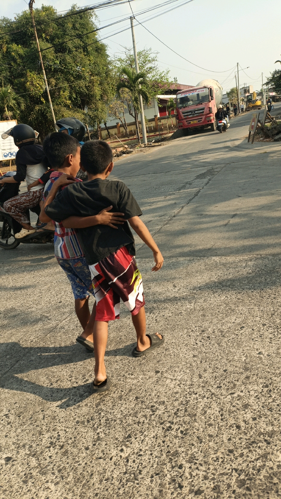 In Jobokuto, Indonesia, two young boys walk hand-in-hand on a paved road. The boy on the left wears a black t-shirt and colorful patterned shorts, while the boy on the right wears a black t-shirt and red, black, and white patterned shorts. They are walking away from the camera, with their backs to the viewer. In the background, a red concrete mixer truck is visible, along with other vehicles and buildings. The sun is low in the sky, casting long shadows. The scene appears to be a typical street in an Indonesian city, with elements of both urban life and a touch of rural charm.