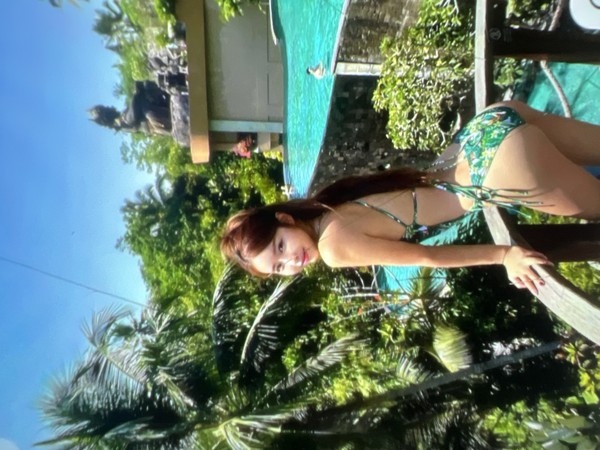 The image shows a woman in a green and white floral bikini posing on a wooden balcony overlooking a tropical landscape. Palm trees are abundant, with lush green foliage filling the background. Beyond the greenery, a bright turquoise swimming pool is visible, with one person swimming in it and another partially submerged. In the far background, a building with a light-colored facade and a statue of a seated figure on its roof can be seen. The sky is a clear blue. The scene appears to be a resort or a similar vacation setting, possibly in a warm, tropical climate. The image does not contain any information or visual cues that would indicate a location in Kara-Dıykan, Kyrgyzstan.
