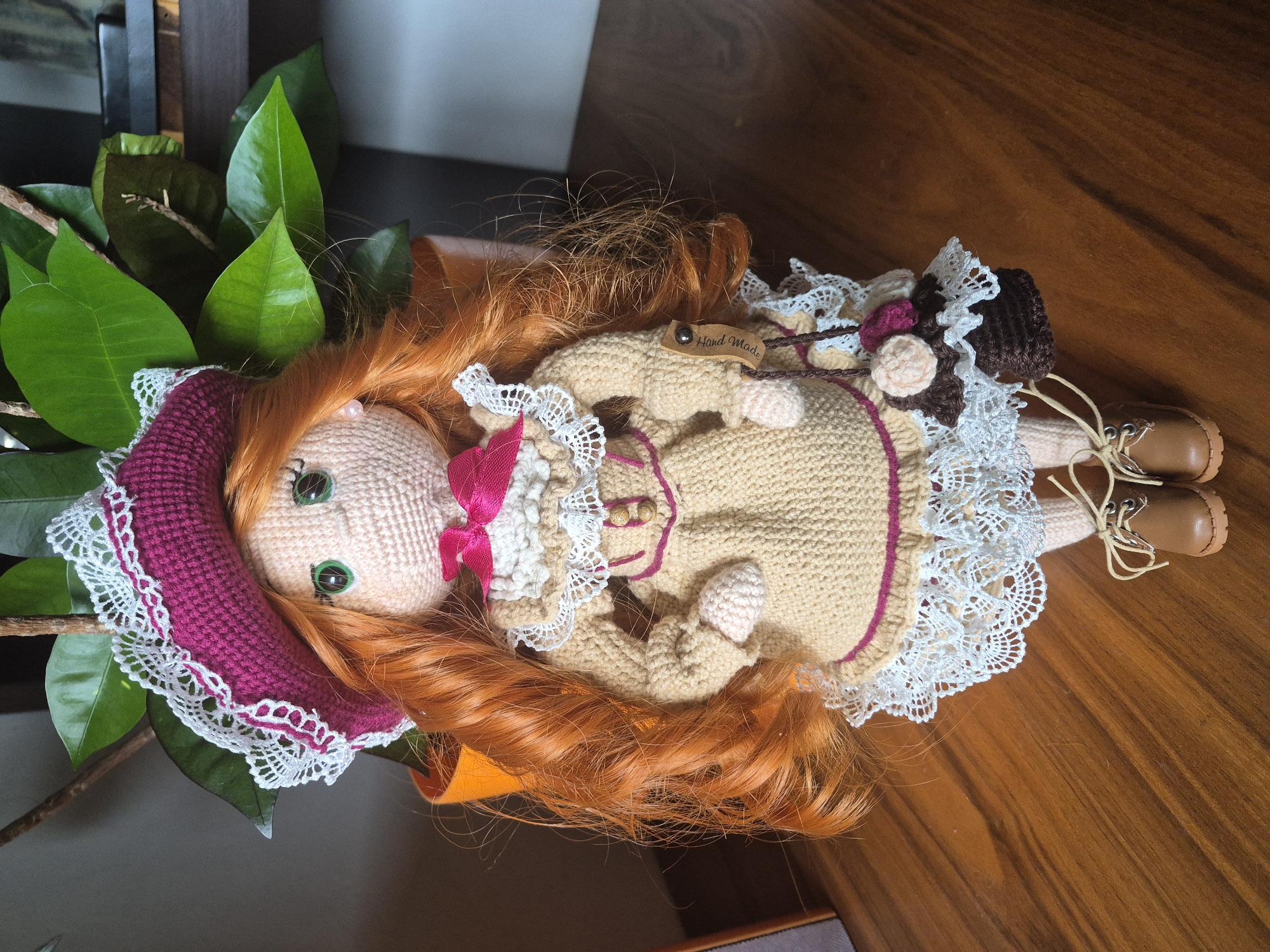 A crocheted doll with auburn hair, green eyes, and a pink bow stands on a wooden surface in front of a plant. The doll is wearing a magenta hat with white lace trim, a beige crocheted dress with pink trim and white lace details, and brown lace-up boots. It is holding a small brown crocheted purse adorned with pink and cream crocheted flowers. A wooden tag on the doll reads "Hand Made". The scene is indoors, likely in a home, as indicated by the wooden table and the plant in a pot. There is no indication of Vlast Truda, Russia in the image.