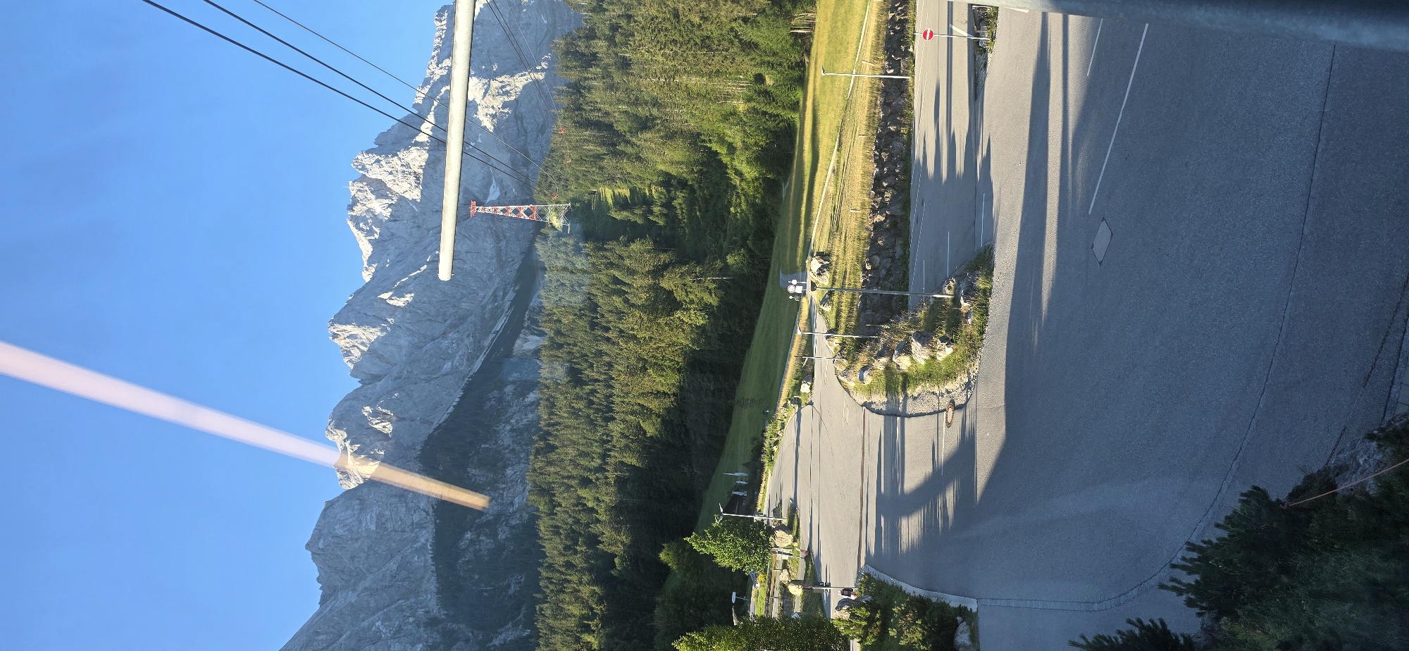 The image captures a view from a cable car ascending towards the Zugspitze mountain in Grainau, Germany. A prominent diagonal beam of light stretches across the clear blue sky from the upper left to the center of the frame. In the background, the majestic, rugged peaks of the Zugspitze mountain range dominate the horizon, their rocky slopes partially obscured by a dense, green coniferous forest that covers the lower slopes. A striking red and white transmission tower is visible amidst the trees, marking the path of a ski lift or cable car line ascending the mountain. In the foreground, a paved road curves through a grassy area dotted with rocks and scattered trees. The road appears to be a parking area or access road, with visible lane markings and shadows cast by unseen objects. The overall scene suggests a sunny day in a mountainous alpine environment.