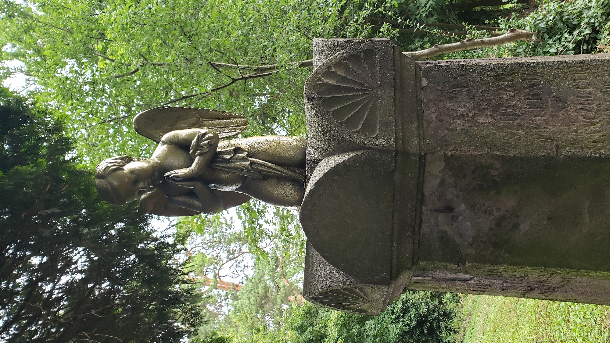 An aged, weathered stone pedestal in a cemetery features a bronze statue of a child angel with folded hands in prayer. The angel has wings and is kneeling on the top of the pedestal. The pedestal itself is octagonal and has decorative carvings of fan-like patterns on its upper surface. The surrounding environment is a lush, green forest with trees and grass. The statue and pedestal appear to be a grave marker. The inscription on the pedestal is illegible due to weathering. This statue is located in the Roman Catholic Cemetery of St. Nicholas in Prešov, Slovakia.