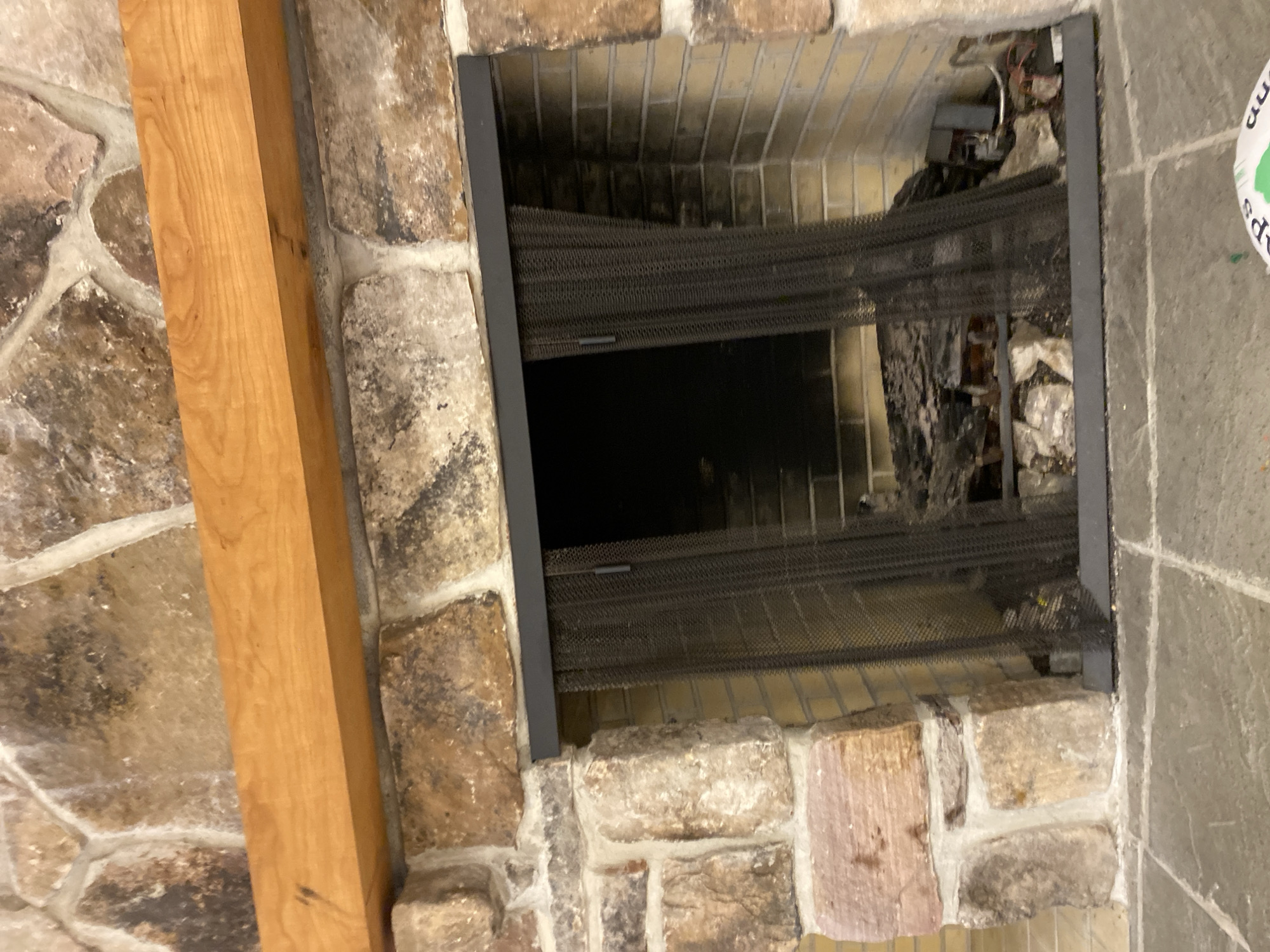 The image shows a fireplace with a stone surround and a wooden mantel. A black mesh screen covers the opening of the fireplace, which contains logs and other debris. The floor in front of the fireplace is tiled. The scene is located in Washington Township, United States of America.