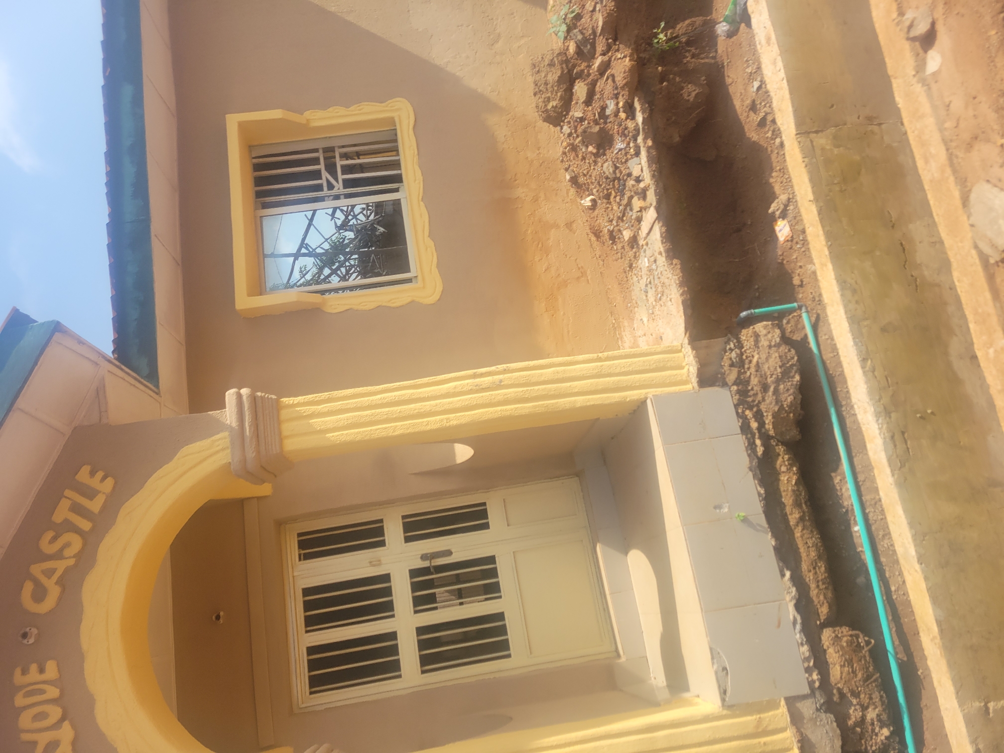 The image shows the exterior of a building, likely a residential property, located in Ogbomosho, Nigeria. The building is painted a light brown color, with yellow accents around the entrance and a window. The entrance features a double door with a metal grate, and two fluted columns flank the doorway, painted yellow. Above the entrance, the words "QODE CASTLE" are visible in raised, yellow lettering. To the right of the entrance, a single window is set into the wall, also with a yellow decorative frame and a metal grate. The window shows reflections of trees and the sky, indicating an outdoor environment.
In front of the building, a section of the ground appears to have been excavated, revealing soil, rocks, and some construction materials. A light blue pipe is visible running along the ground in the excavated area. The overall scene suggests a property that is either under construction or undergoing some form of renovation. The lighting suggests it is daytime.