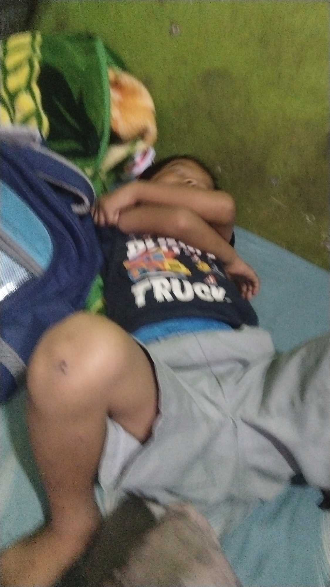 A young child is asleep on a light blue mattress, positioned on their back with their arms crossed over their chest. The child is wearing a dark blue t-shirt with a graphic of a truck and the word "TRUCK" in white lettering, and light gray shorts. The child's left leg is bent at the knee, with a small mark visible on the kneecap. To the left of the child is a dark blue backpack. Behind the child, a green blanket with a yellow and green pattern is partially visible, along with an orange, fluffy object. The background consists of a textured green surface, possibly a wall or carpet, with some shadows. The scene appears to be indoors.