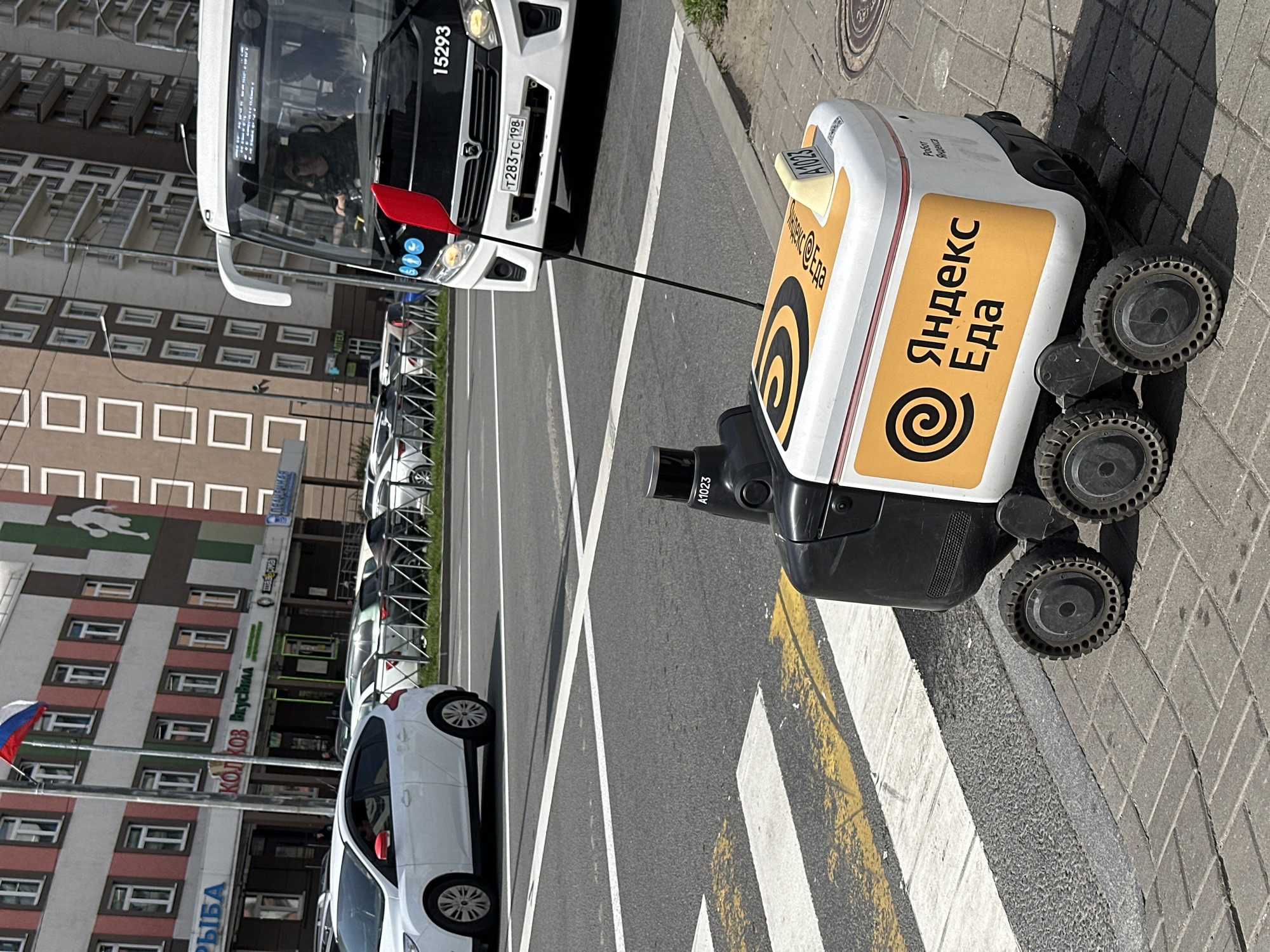 Here is a description of the image:
The image depicts a scene in Murino, Russia, showing a Yandex.Eda delivery robot near a crosswalk, with a bus and a white car nearby. The robot, which is primarily white with yellow accents and the Yandex.Eda logo, stands on the sidewalk near the crosswalk. It has six wheels and a camera visible on the front. A black cable is connecting it with the bus.
A white bus is positioned on the road with the license plate number "T283TC 198" visible. The number "15293" is also visible on the upper portion of the bus. In front of the bus there is a red flag.
A white car is parked along the road, and several other cars are visible in the background. Across the road are buildings with multiple stories and windows. Signage on the buildings include "РЫБА" (Fish), "Вкусна" (Delicious), "КОЛ КОЗ" (Dairy Product).