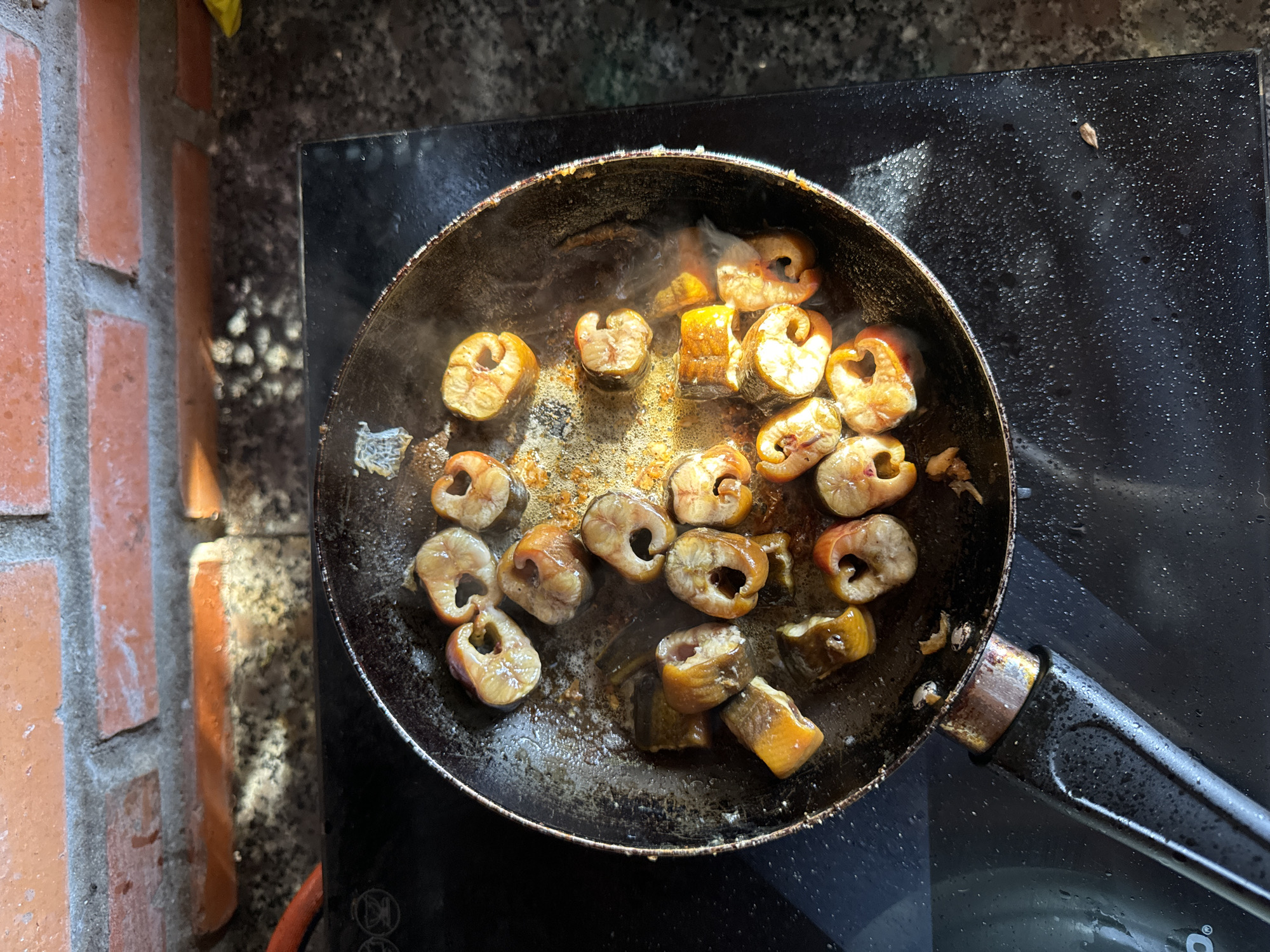 A top-down shot captures a frying pan on a black stovetop. The pan contains segments of what appears to be eel, cooking in oil or fat. Steam rises from the food. The stovetop is slightly wet. The scene is partially lit by natural light coming from the left, illuminating a section of a brick wall. The setting suggests an outdoor cooking area. Without further context, it is not possible to confirm this image was taken in Mũi Né, Vietnam.