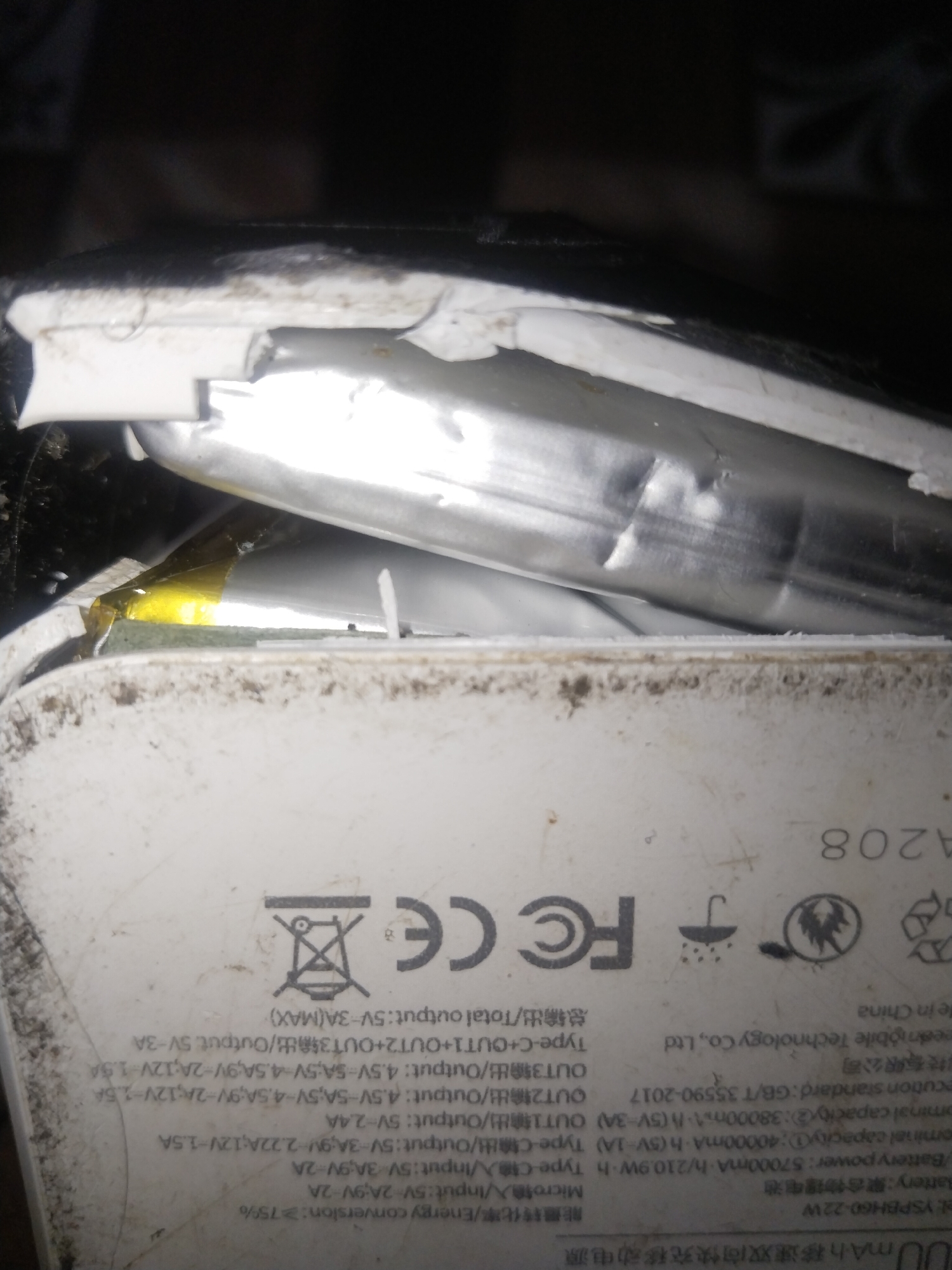 The image shows a damaged portable power bank. The white plastic casing is broken, revealing the internal battery components which appear to be swollen and metallic. The device is dirty and dusty, suggesting it may have been exposed to harsh environmental conditions. Text and symbols are visible on the casing, including "FC", "CE", a recycling symbol, and information about battery capacity and output ports. The text also includes "YSPBH60-22W". The location in Uyo, Nigeria, is not discernible from the image itself.