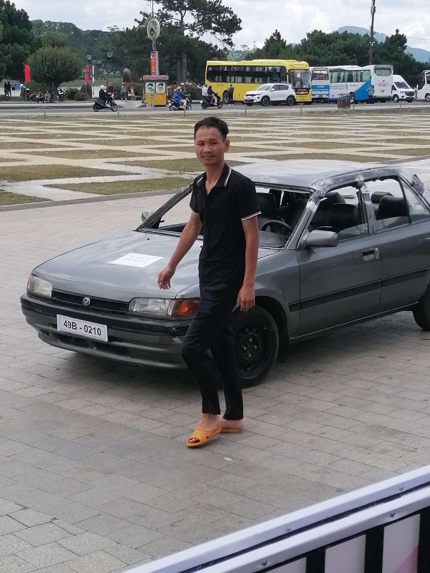 A man in a black polo shirt and black pants stands to the right of a gray hatchback car. The car's hood is slightly open, and a white piece of paper is placed on it. The license plate reads "49B - 0210." The man is wearing orange flip-flops. The scene appears to be an outdoor plaza with patterned concrete paving. In the background, there are trees, several buses, and a white van parked in a line. The sky is overcast. This location is likely the central plaza or a public space in Da Lat, Vietnam, known for its parks and public gathering areas.