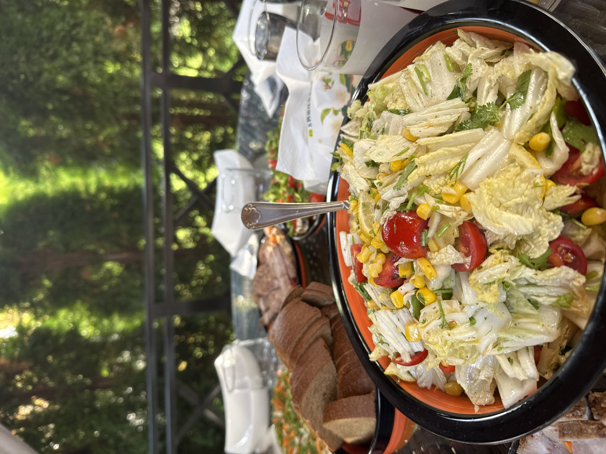 An outdoor dining scene, likely in Sary-Oy, Kyrgyzstan, features a spread of food on a table. The foreground is dominated by a large bowl of salad, predominantly composed of chopped napa cabbage, cherry tomatoes, and corn kernels. A fork is partially submerged in the salad. To the left of the salad bowl is a platter of sliced brown bread. In the background, other food items and drinks are visible, including what appears to be a pizza and some sliced meats. The setting is outdoors with lush green trees and foliage visible behind a dark railing, suggesting a garden or patio environment. The lighting indicates it is daytime. There are no people clearly visible in the image.
