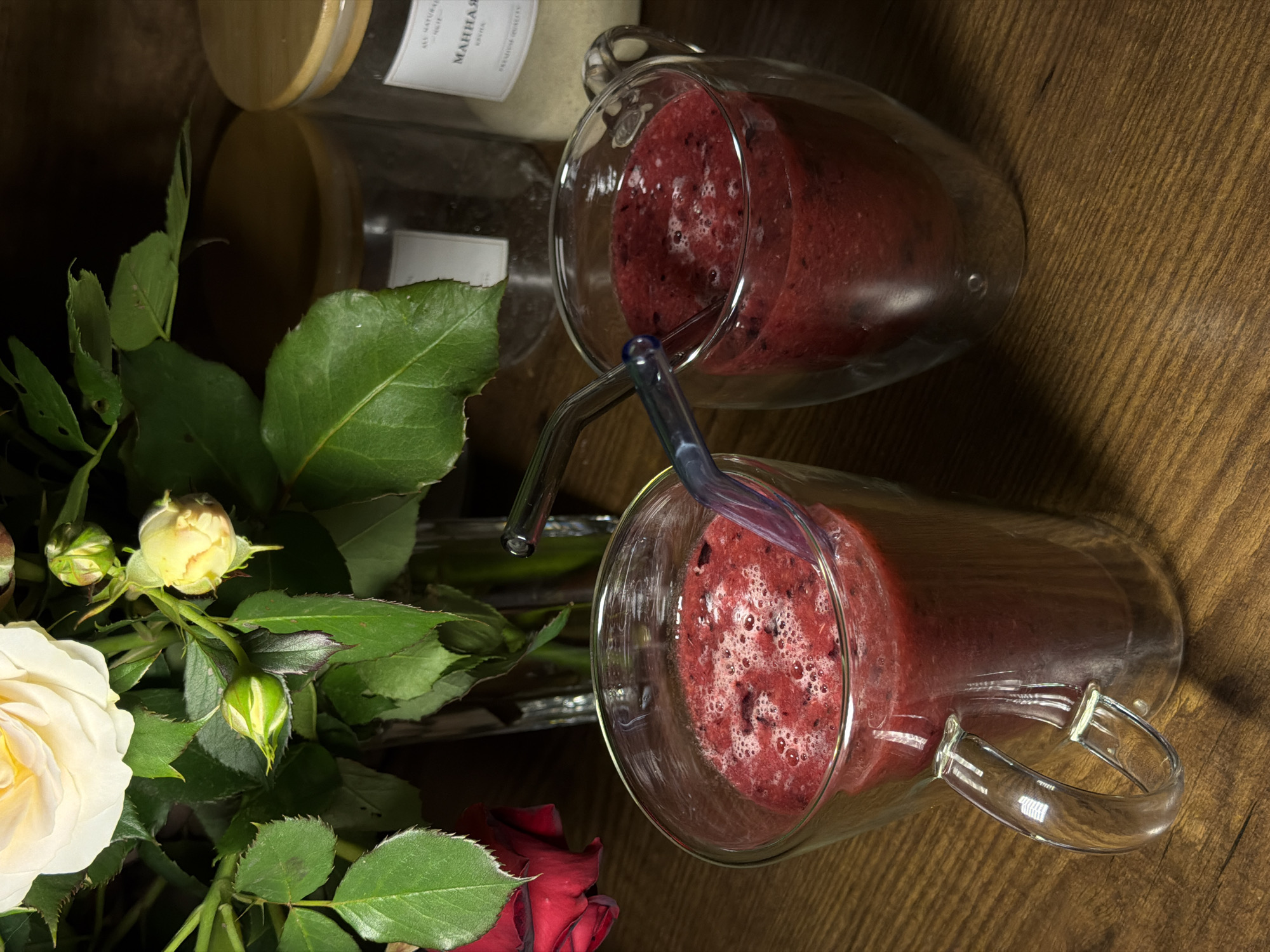 Two glass mugs filled with a red beverage, each with a bent glass straw, sit on a wooden surface. Adjacent to them are glass jars, one with a wooden lid. A bouquet of roses in shades of white, yellow, and red is situated next to the mugs. The scene appears to be indoors, possibly in the city of Rossiyskiy, Russia, though this location is speculative. There are no people visible in the image.