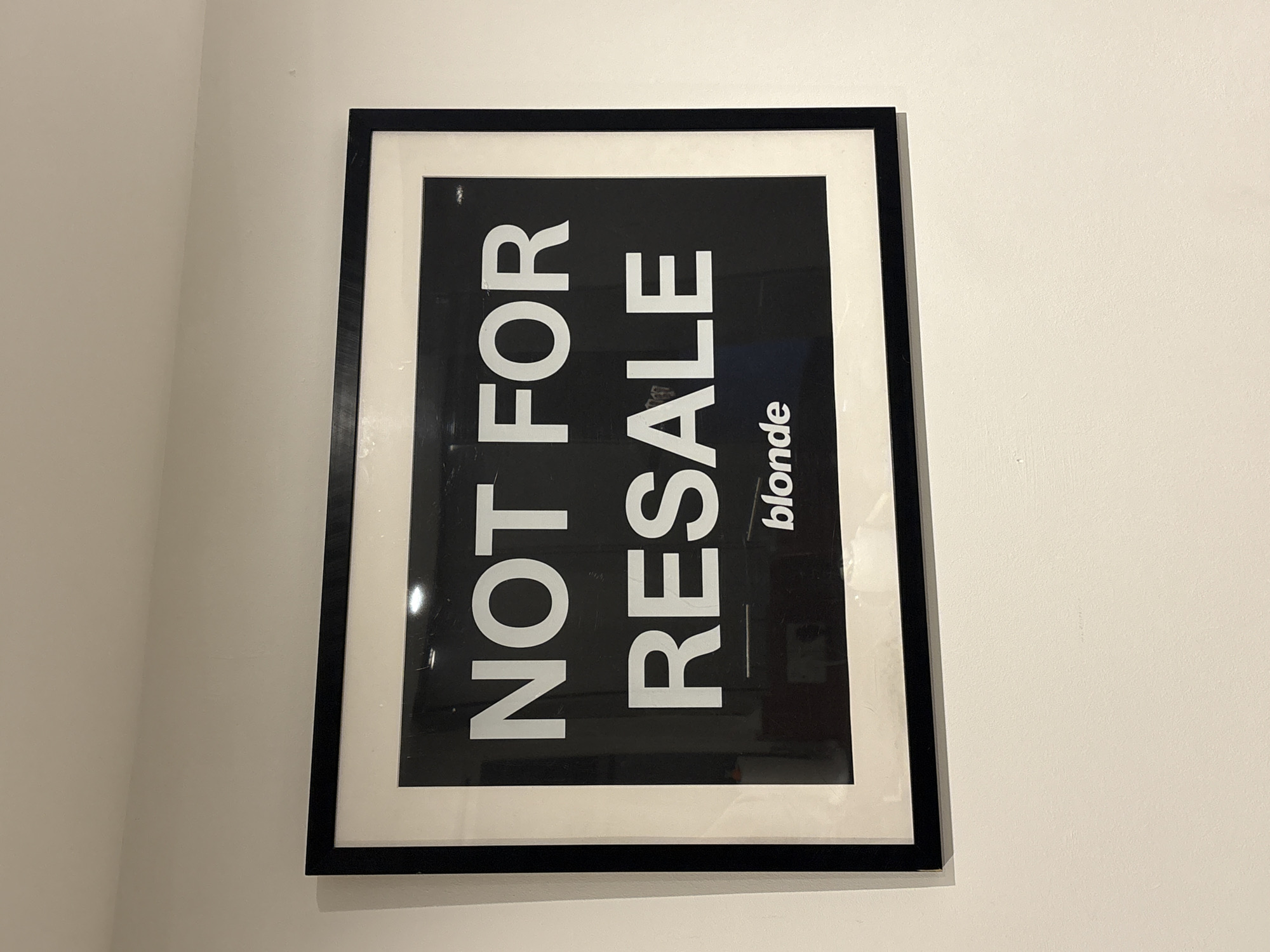 The image displays an indoor scene in the city of London, United Kingdom. A rectangular framed poster is centrally mounted on a plain, light-colored, possibly off-white or cream, painted wall. The poster features a black background with prominent white sans-serif text. The top line reads "NOT FOR", and the second line reads "RESALE". Below this, centered near the bottom, is the word "blonde" in a smaller, white sans-serif font. The poster is enclosed within a black frame, which surrounds a white mat border that separates the artwork from the frame's edge. The protective glass covering the poster exhibits reflections of the ambient lighting and potentially other room elements, along with minor surface dust or scuffs. No people, specific actions, or other objects are discernible in the immediate environment. The lighting appears to be artificial interior illumination. No specific architectural details or external views are present to indicate a precise location within London.