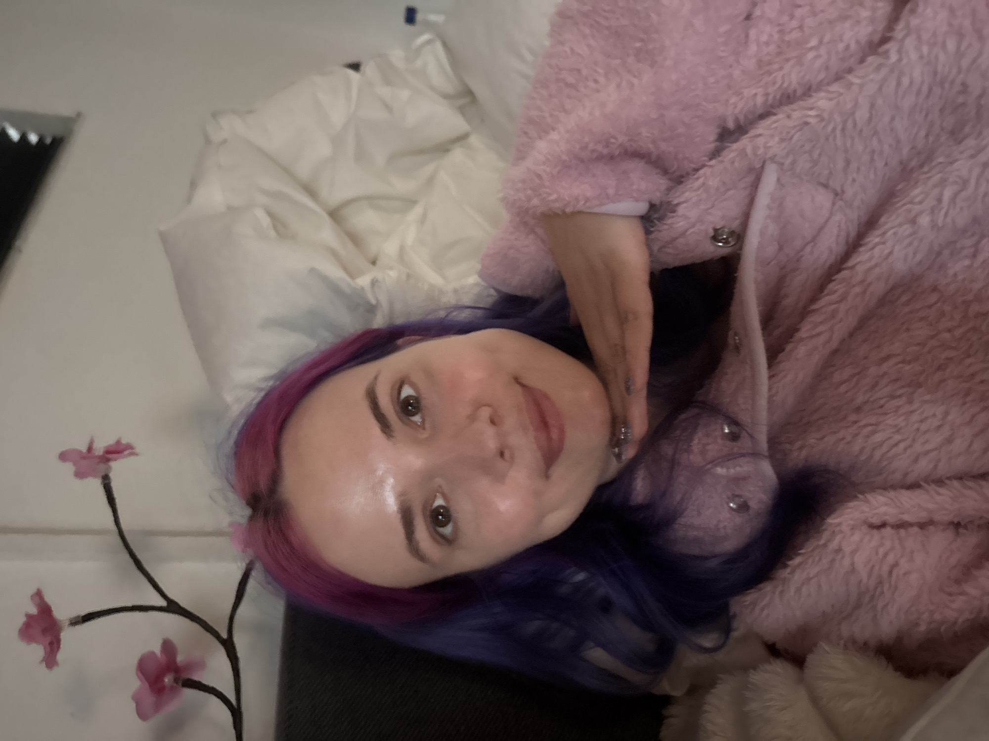 A young woman lies on a bed. Her hair is dyed purple and pink. She is wearing a pink, fluffy jacket. There is a white comforter behind her. A faux flower branch is in the foreground. The location is not discernible.