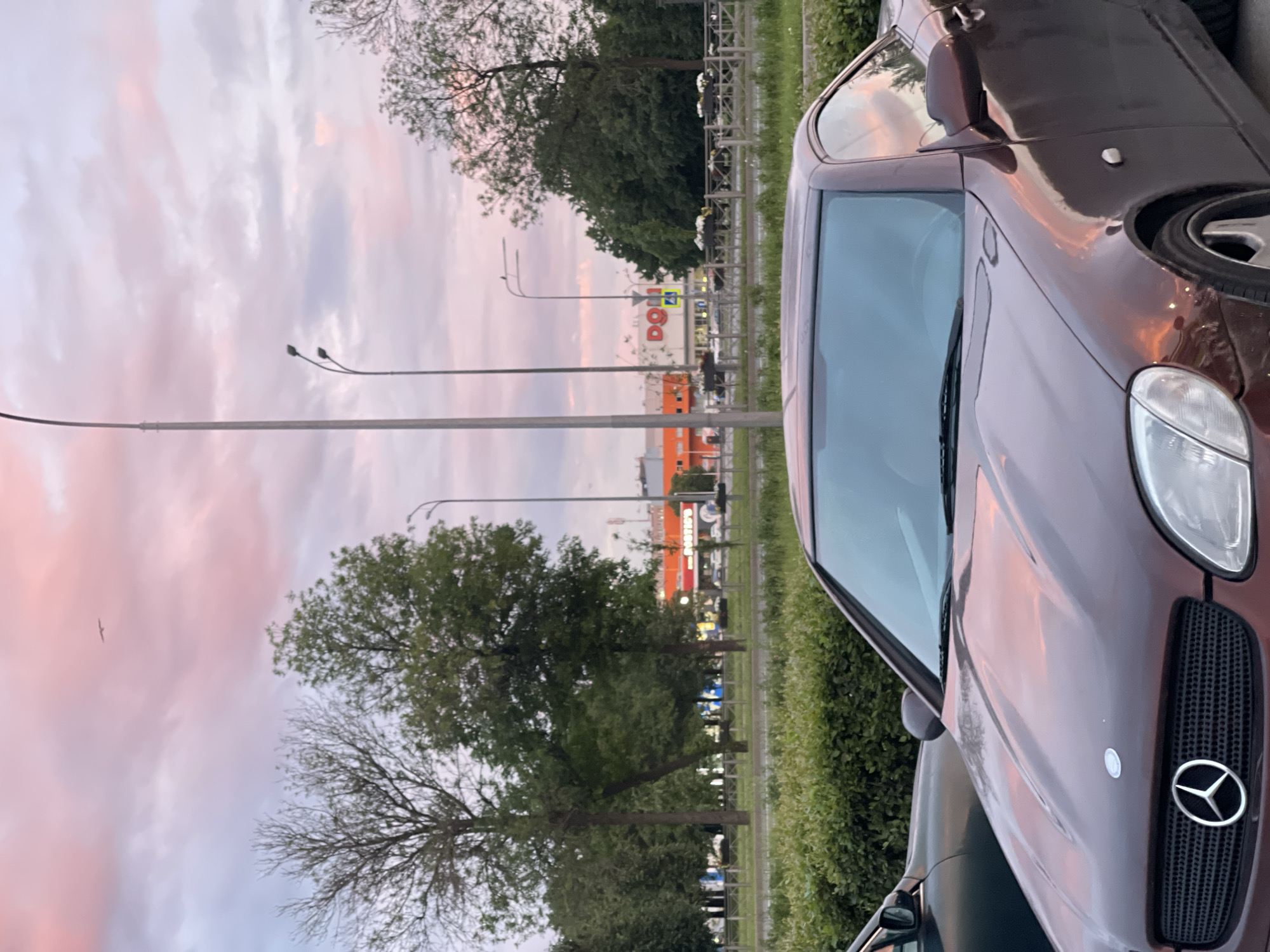 The image shows a street scene with a maroon Mercedes-Benz convertible in the foreground. The car is parked on pavement with a hedge and trees behind it. In the background, there is a large orange building with the word "DONS" visible, likely a store or shopping center. The sky is a soft pink and purple, indicating sunset or sunrise. Several streetlights are visible, along with trees and other vehicles on the road. A single bird is flying in the sky. The overall scene appears to be an urban environment during twilight.