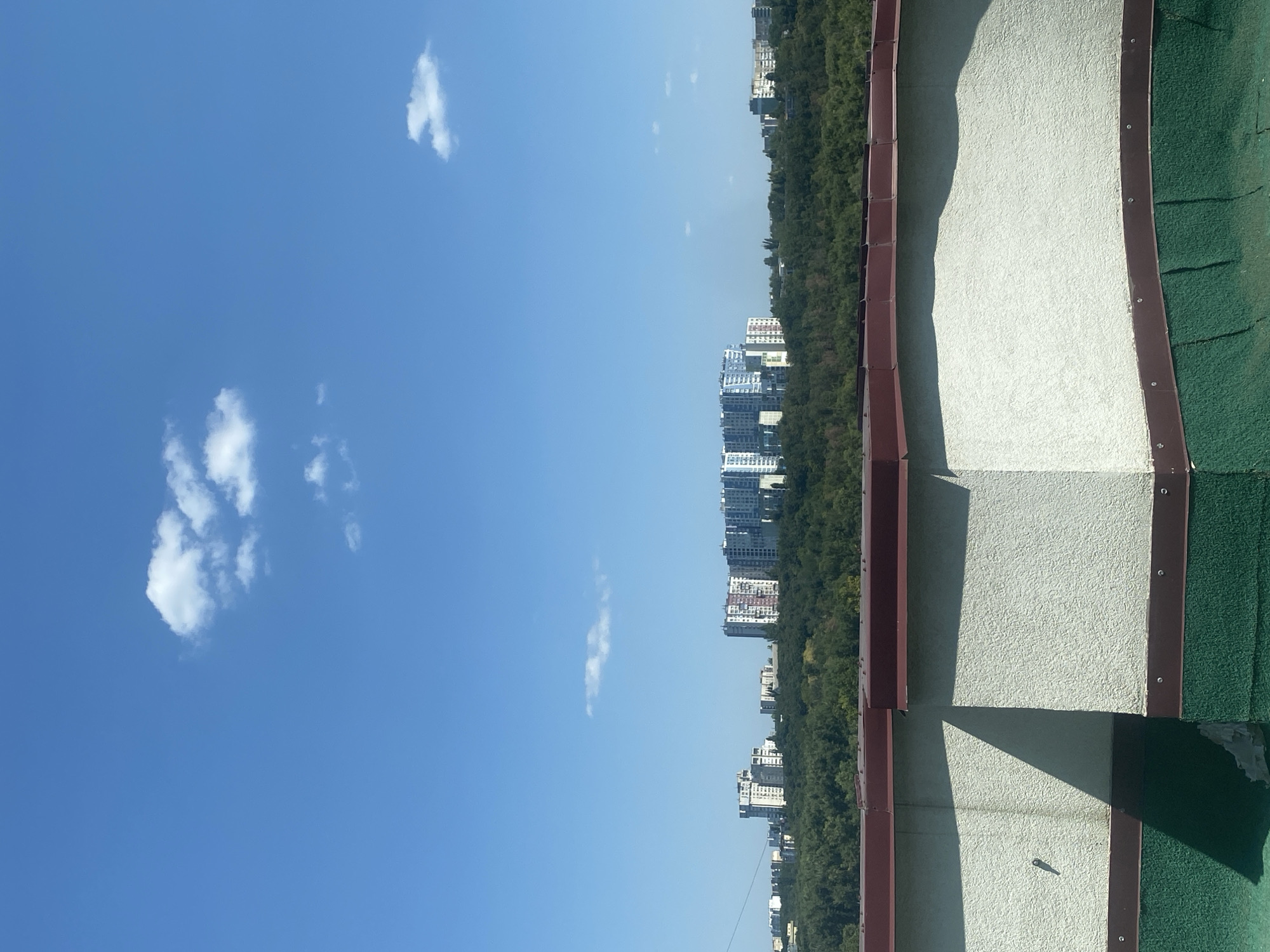 An outdoor, elevated shot features a clear blue sky with scattered white clouds. In the middle ground, a dense, green forest line separates the foreground from a cityscape of apartment buildings. The apartment buildings are modern, with varying heights and architectural styles, appearing to be in a residential area. The foreground is dominated by a roof or elevated platform with a textured, light-colored wall and a dark green lower section, bordered by a dark red trim. The scene appears to be a sunny day in Chișinău, Moldova, likely a residential district with a significant green space. There are no people or animals visible in the image.