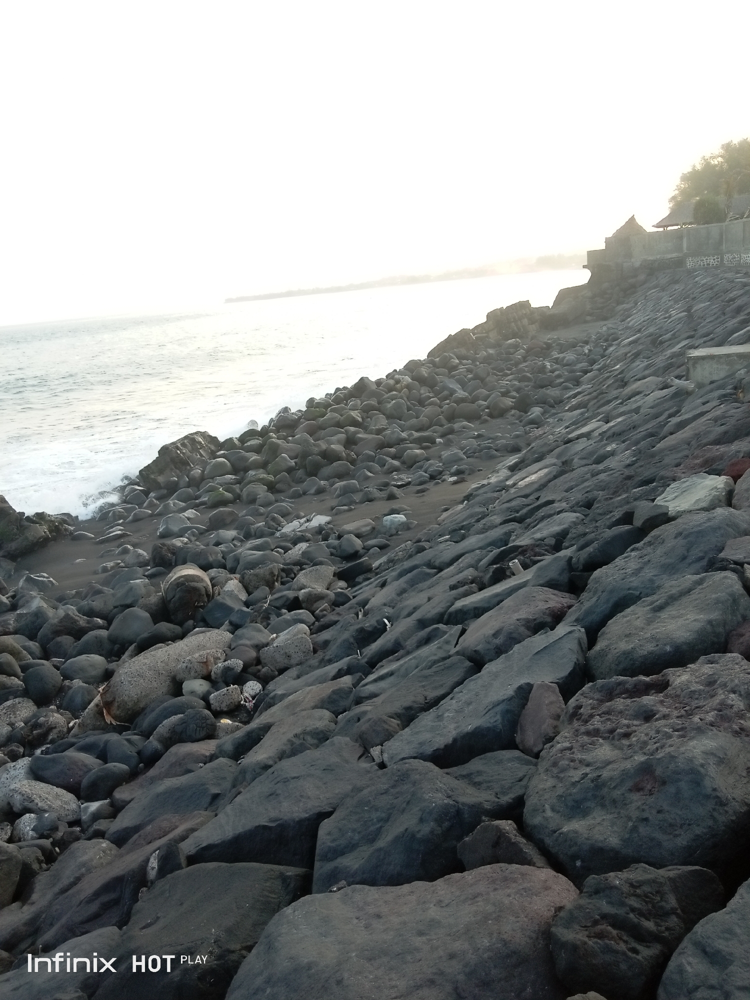 The image depicts a rocky shoreline with the ocean in the background. The foreground is dominated by a dense collection of dark, irregularly shaped rocks and boulders of varying sizes, some appearing smooth and water-worn, others rough and angular. A narrow strip of black sand beach is visible to the left, with gentle waves breaking. In the distance, the horizon is hazy and bright, suggesting a sunset or sunrise. A concrete structure with thatched-roof huts or shelters is perched on a higher elevation behind the rocks on the right side of the image. The water is a muted grey-blue. The scene is devoid of people. The Infinix HOT PLAY watermark is present in the bottom left corner. This scene is likely located along the coast of Banjarangkan, Indonesia, a region known for its volcanic rocky shorelines.