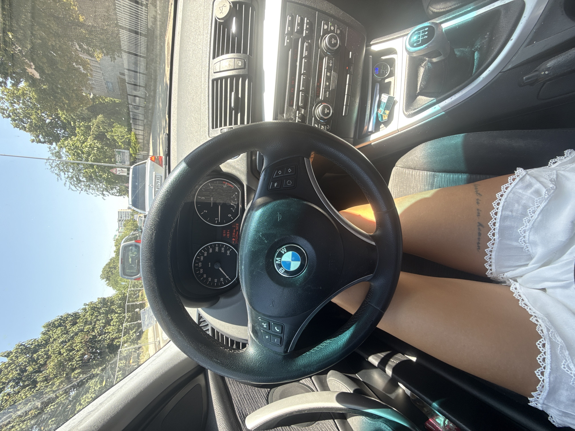 The image is taken from the interior of a BMW vehicle, likely on a sunny day in Plovdiv, Bulgaria. The perspective is from the driver's seat, focusing on the steering wheel, dashboard, and the driver's legs. The steering wheel prominently displays the BMW logo. The dashboard shows the car's instrument cluster with a speedometer and tachometer, as well as the car's radio and air conditioning controls. A manual gear shifter is visible in the center console. The driver's legs are visible in the lower part of the frame, wearing a white lacy garment. Outside the car, trees and other vehicles can be seen through the windshield, suggesting the car is in traffic or parked on a street with vegetation. The overall impression is a snapshot of a person in their car, possibly during a commute or a drive through the city.