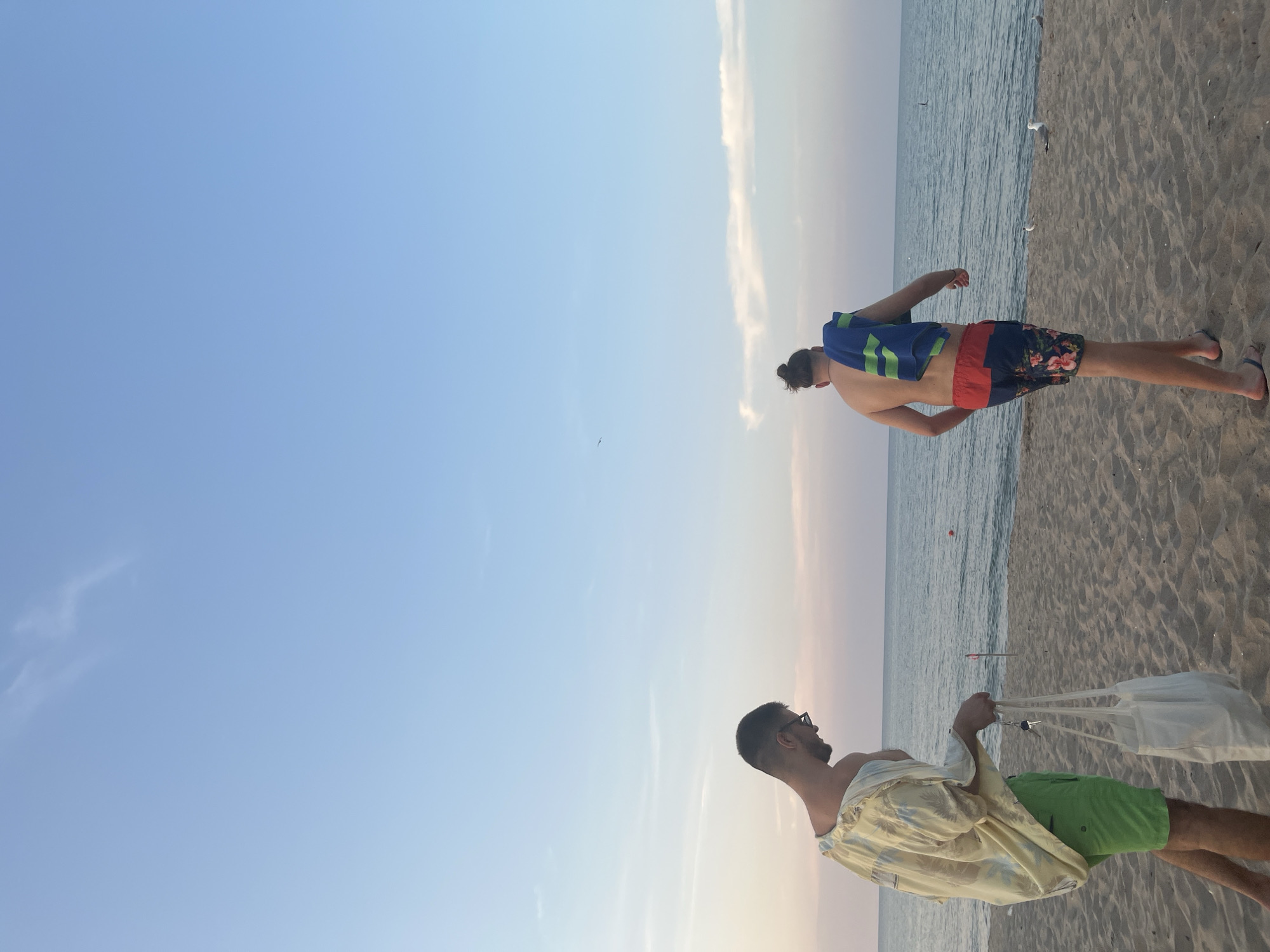 Two men are walking on a sandy beach by the sea. The man on the left is wearing a yellow, loosely fitting shirt and green shorts, and is carrying a white tote bag and keys. He is looking down. The man on the right is facing away from the camera, wearing red and blue patterned swim shorts, with a blue and green towel draped over his shoulder. He is walking towards the sea. The sky is a pale blue with wispy clouds, and the sea is calm. The overall impression is of a relaxed summer day at the beach. There are no visible landmarks that would place this specific scene in Lozenets, Bulgaria.
