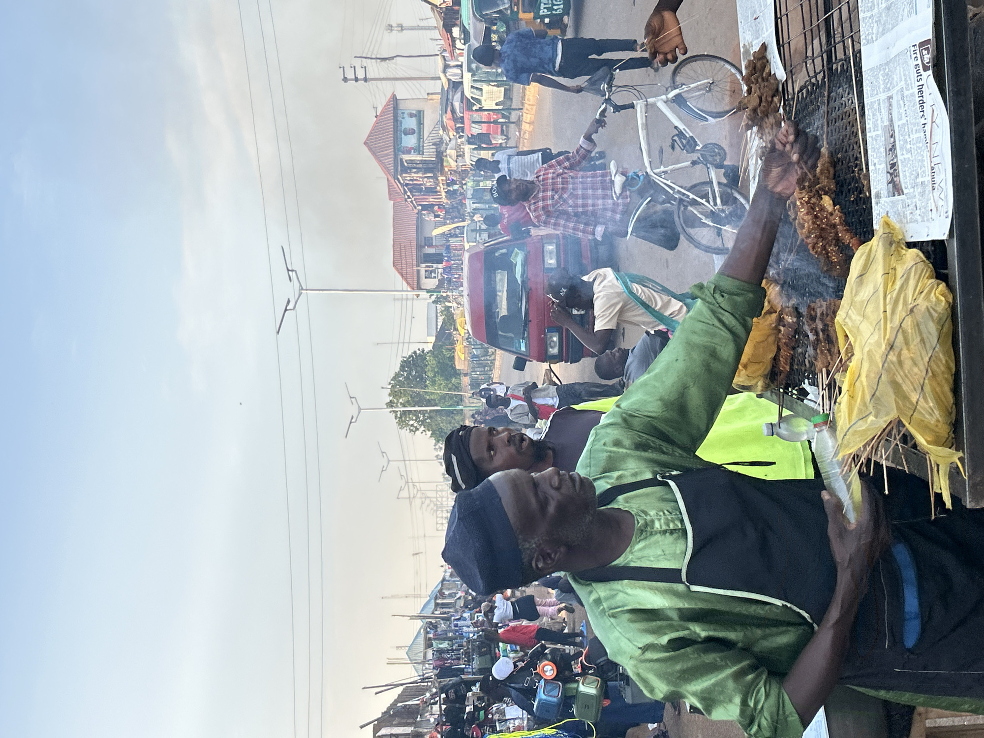 A bustling street scene in Jos, Nigeria, shows a man in a light green shirt and black apron grilling meat skewers on a charcoal grill, creating smoke that fills the air. The grill is backed by newspaper and has yellow bags hanging near it. Several people stand nearby, and a man on a bicycle is visible behind the grill. In the background, there are vehicles, including a red van, and numerous pedestrians moving about, indicating a crowded market or commercial area. Power lines are visible overhead. The sky is slightly overcast.
