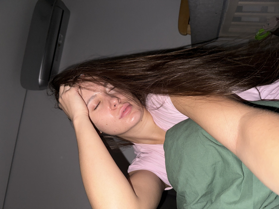 A female subject, appearing to be a young adult, is depicted in a close-up, chest-up shot, indoors in Molodi, Russia. Her eyes are closed, and her head is slightly tilted to the right. Her long, straight, dark brown hair falls over her right shoulder and partially obscures her face. Her right hand is raised, with fingers touching her forehead and running through her hair. Her skin exhibits a sheen, particularly on her forehead, nose, and lips, which are slightly parted. She is wearing a light pink, short-sleeved garment, possibly ribbed. A dark green fabric, consistent with bedding or a blanket, covers her left arm and torso. The background is dimly lit, revealing a grey wall. A light-colored, wall-mounted air conditioning unit is visible in the upper right quadrant of the image. A portion of a light-colored wooden structure, possibly a crib or bed frame with vertical slats, is present in the lower right background.