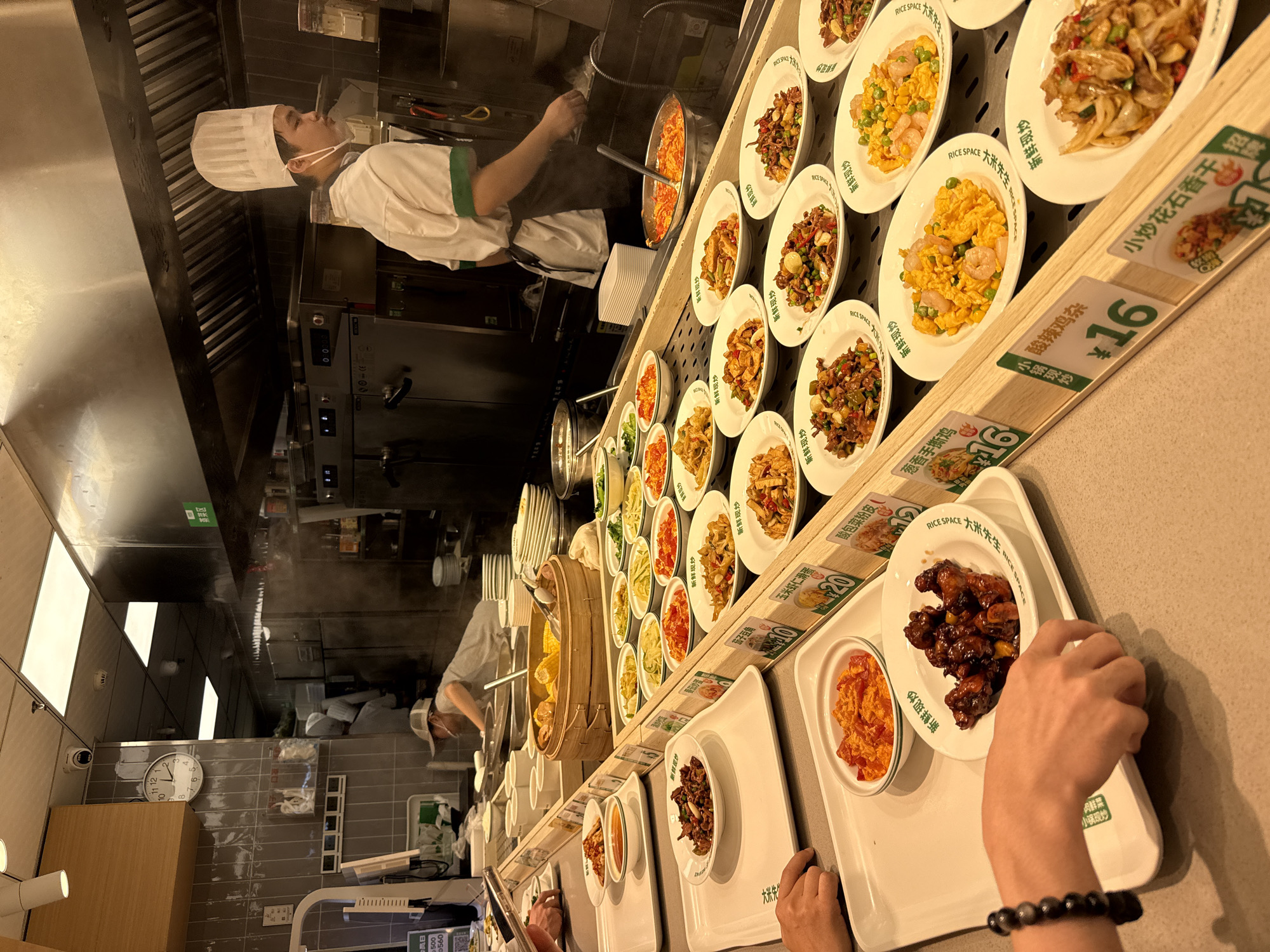 A self-service food buffet with a variety of Chinese dishes is presented. In the background, a chef in a white uniform and hat is visible, working in the kitchen area. In the foreground, customers are selecting food items. The buffet line features numerous white trays and bowls filled with different prepared foods, including stir-fried vegetables, meats, and seafood. Signs with prices and dish names in Chinese are placed along the buffet line. The environment appears to be an indoor establishment, likely a restaurant or food court, in Tianhe District, China. The lighting is bright, typical of a commercial food service setting.