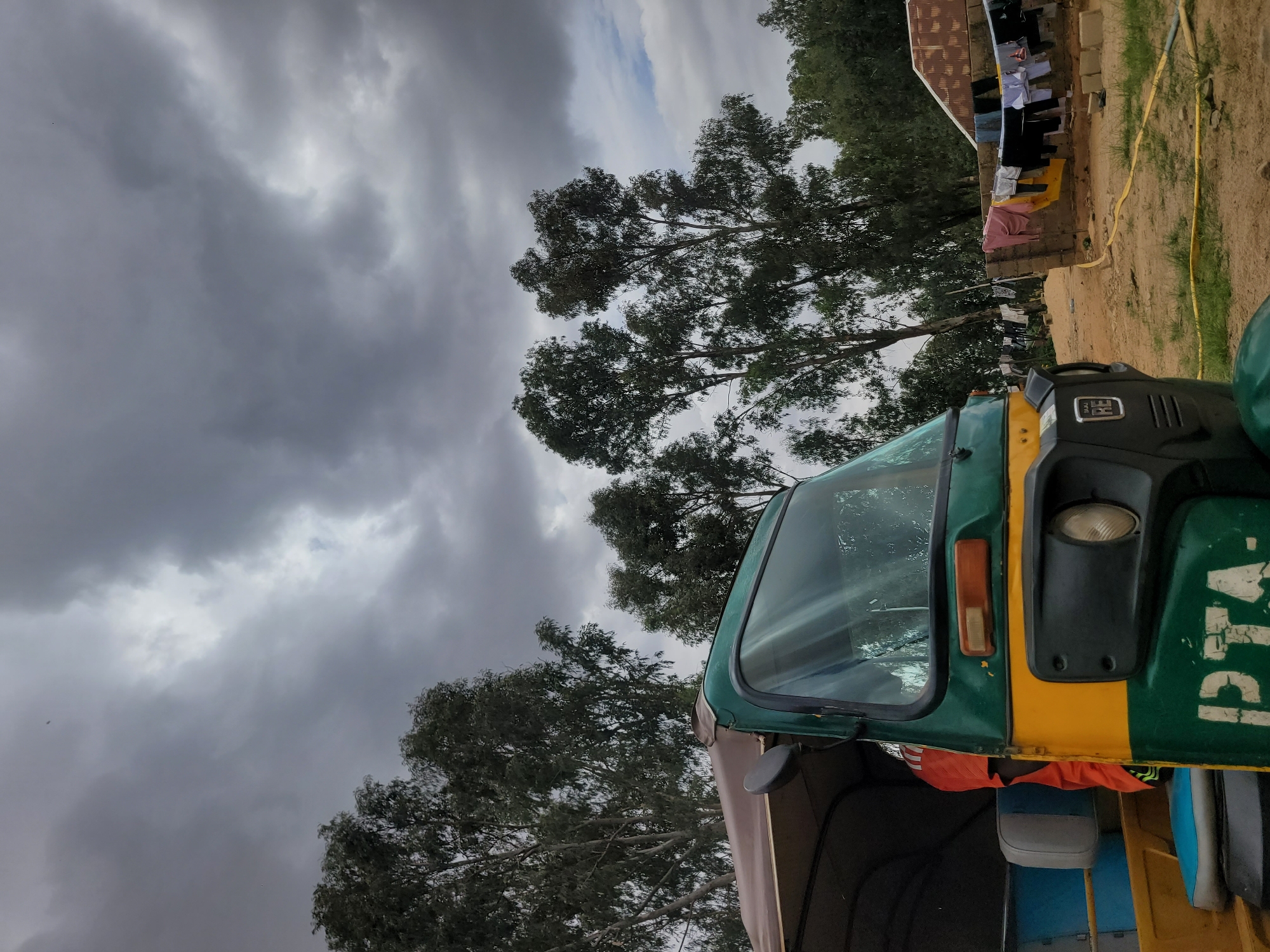 This outdoor image, taken in Jos, Nigeria, captures a static scene under a heavily overcast sky, suggesting an impending rain or dull weather. The dominant subject is a green and yellow three-wheeled auto-rickshaw, partially visible on the right side of the frame. Its front windshield, a single headlight, and an orange indicator light are clearly seen. Text, partially obscured and worn, resembling "PTA" is visible on the green bodywork, and a small "RE" emblem is present on a black panel above the headlight. Inside the auto-rickshaw, a blue and a grey seat are discernible, along with some orange fabric hanging below.
The background is dominated by numerous tall trees with dense green foliage, set against a dramatic sky filled with dark, heavy clouds. To the far right, a simple, low-rise building with a reddish-brown roof is visible, outside of which various items of colorful laundry are hung on a clothesline to dry. The ground is unpaved, composed of reddish-brown dirt with patches of green grass, and a yellow hose or rope lies coiled on the ground.
The scene depicts a quiet moment in a residential or semi-urban environment, with the parked vehicle and drying clothes indicating human activity, though no people are visible. The heavy cloud cover suggests daytime, possibly morning or afternoon, but without direct sunlight.