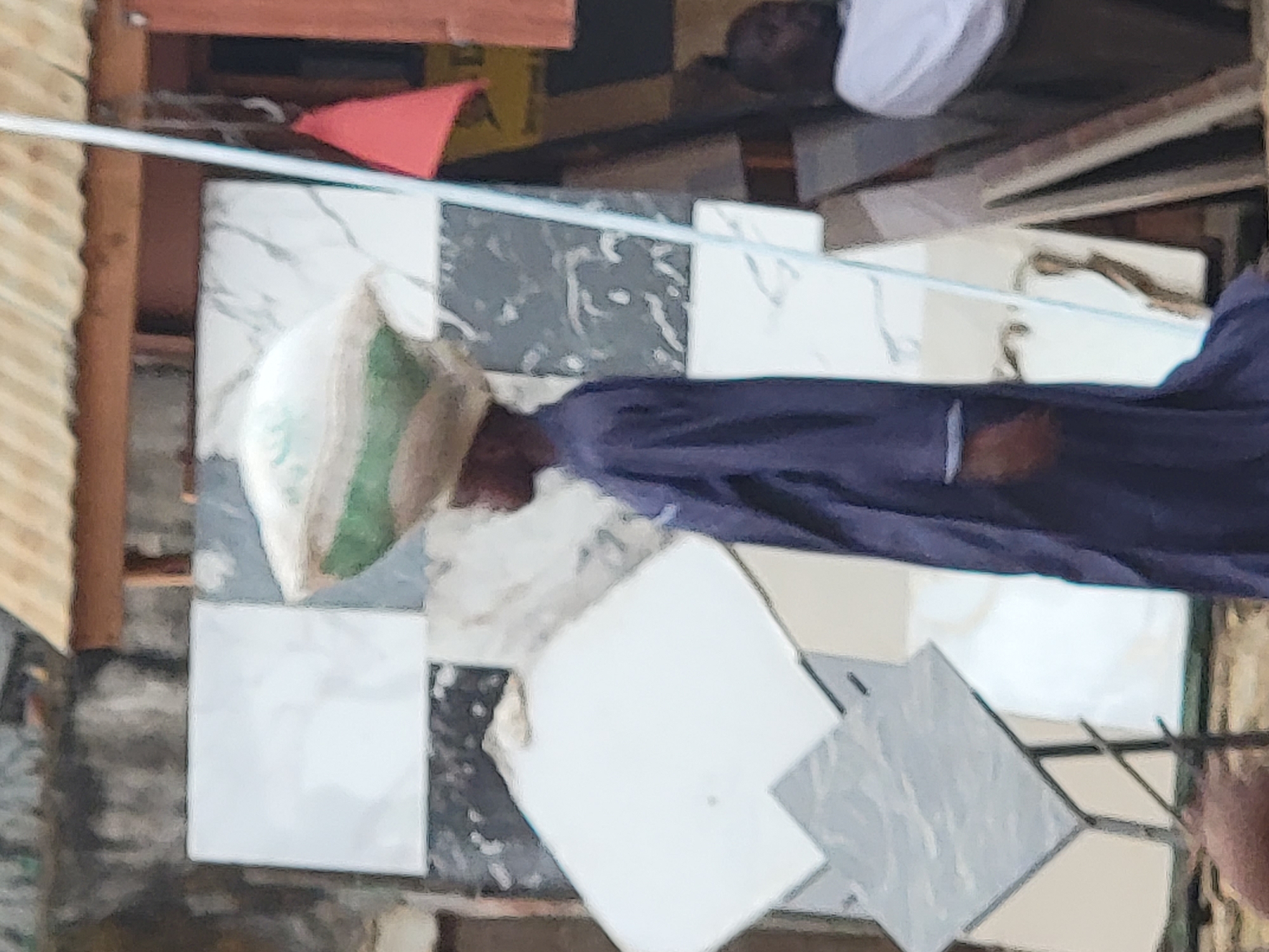 This image depicts an outdoor market or roadside stall scene in Maiburiji, Nigeria, during daylight hours. A person is centrally positioned, standing and viewed from the side, wearing a dark blue, long-sleeved garment. They are carrying a light-colored sack, possibly containing goods, balanced on their head. Their gaze appears directed downwards.
The foreground and mid-ground prominently feature a collection of various ceramic tiles, arranged on what appears to be a display surface or the ground. These tiles exhibit different designs, including solid white, white with distinct gray marble-like veins, and dark gray with white specks or patterns. A thin, light-colored rope or cord stretches horizontally across the mid-section of the image, above the tiles.
The background shows elements of makeshift construction, including wooden beams and a corrugated metal sheet on the far left, suggesting a stall or building structure. A small red triangular object is visible in the upper left background. Another individual, dressed in a white garment, is partially seen in the upper right background. The scene suggests a commercial setting, likely focused on the sale or display of building materials like tiles. No discernible text is visible.