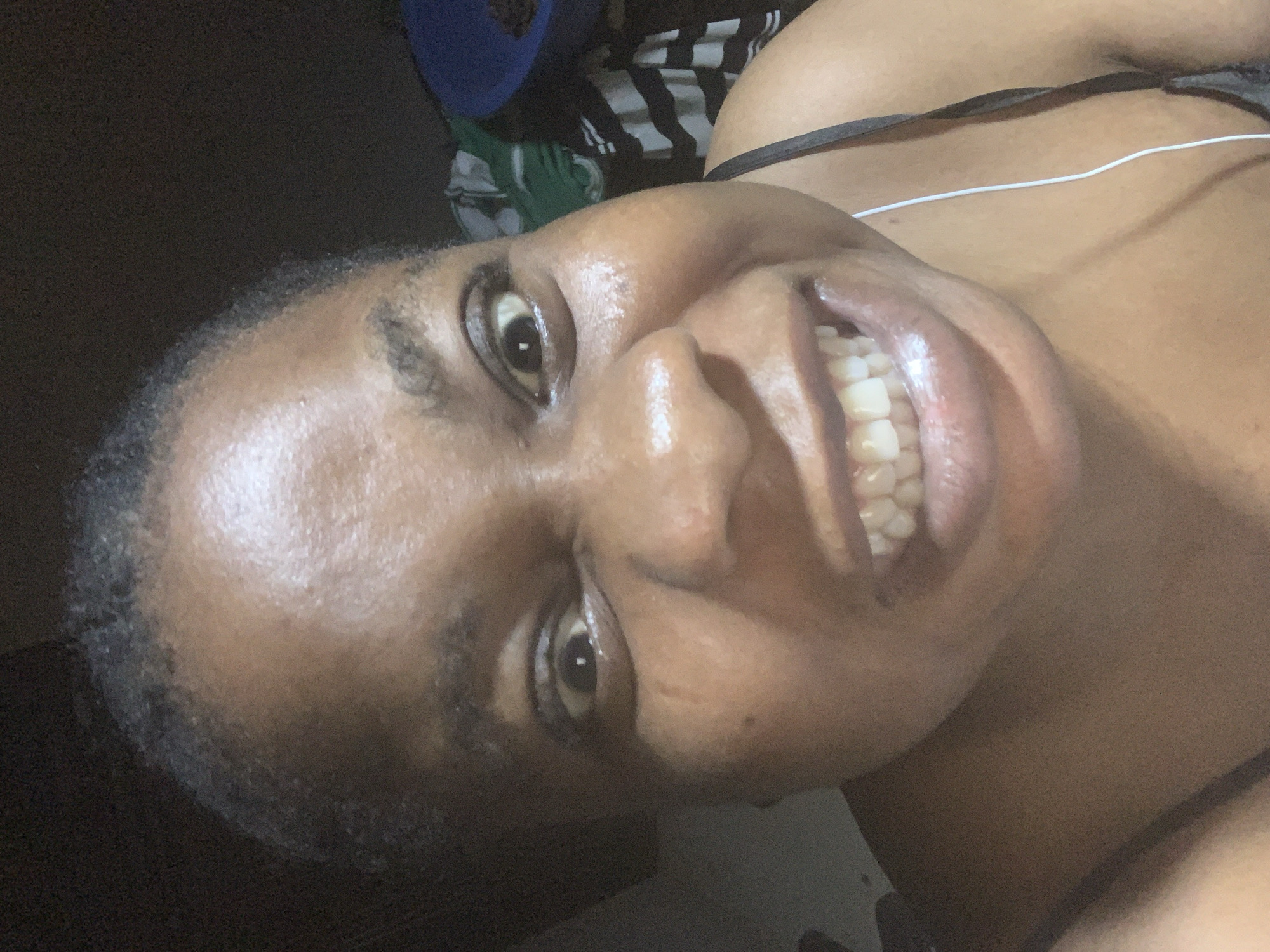 A close-up, angled shot shows a Black woman with dark hair smiling. Her teeth are visible. She is wearing a thin-strapped top, and a white wire is visible near her neck. The background is dark and indistinct, showing some clothing items. The city and exact location are unknown.