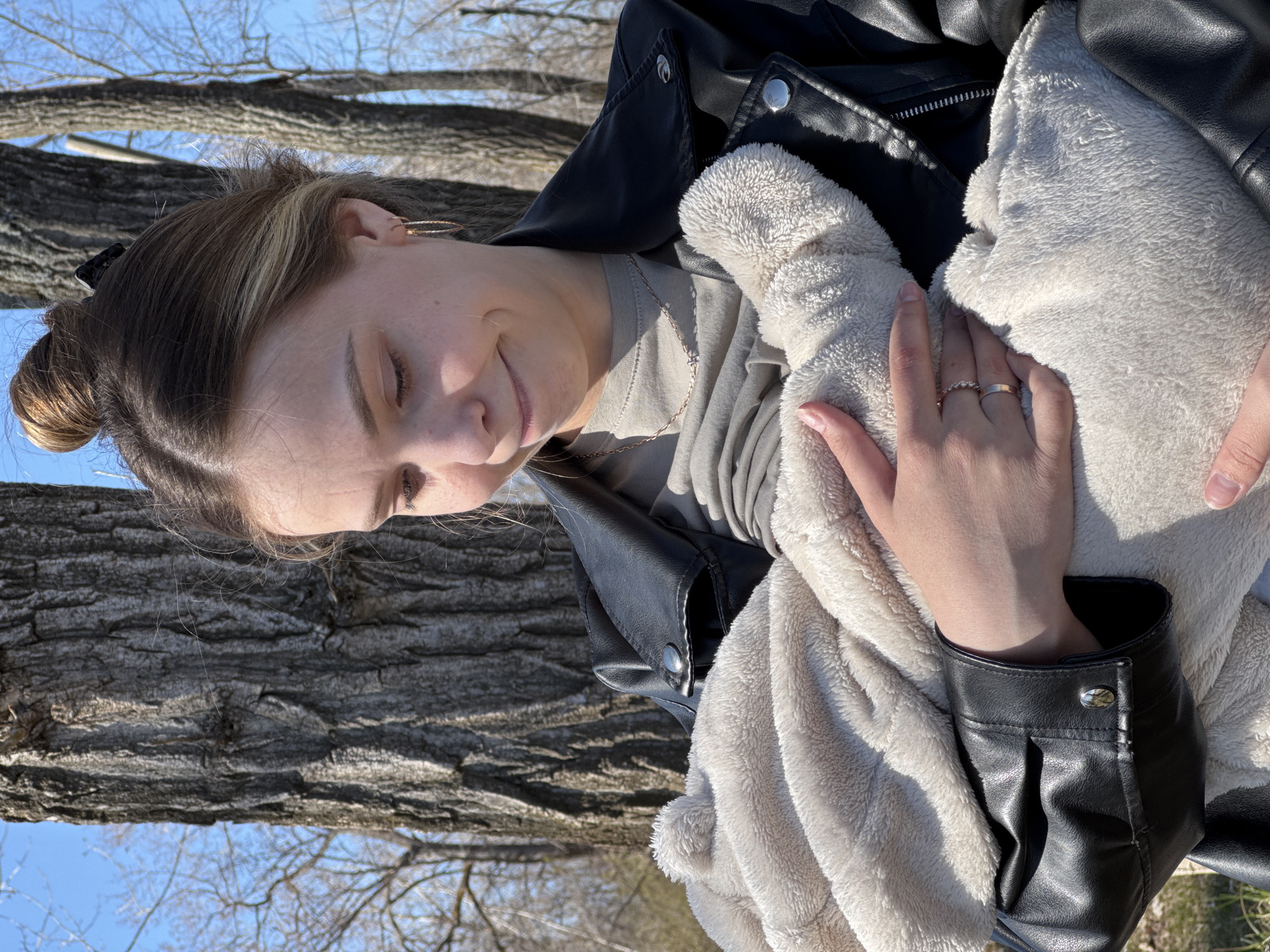 A woman is seen in an outdoor setting. She has light brown hair pulled back into a bun with blonde highlights. She is wearing a light grey t-shirt and a black leather jacket. In her arms, she holds a soft, light-colored blanket. The background suggests a park-like environment with a tree trunk visible behind the woman and blue sky with bare tree branches above. The location is Chaikovsky, Russia.