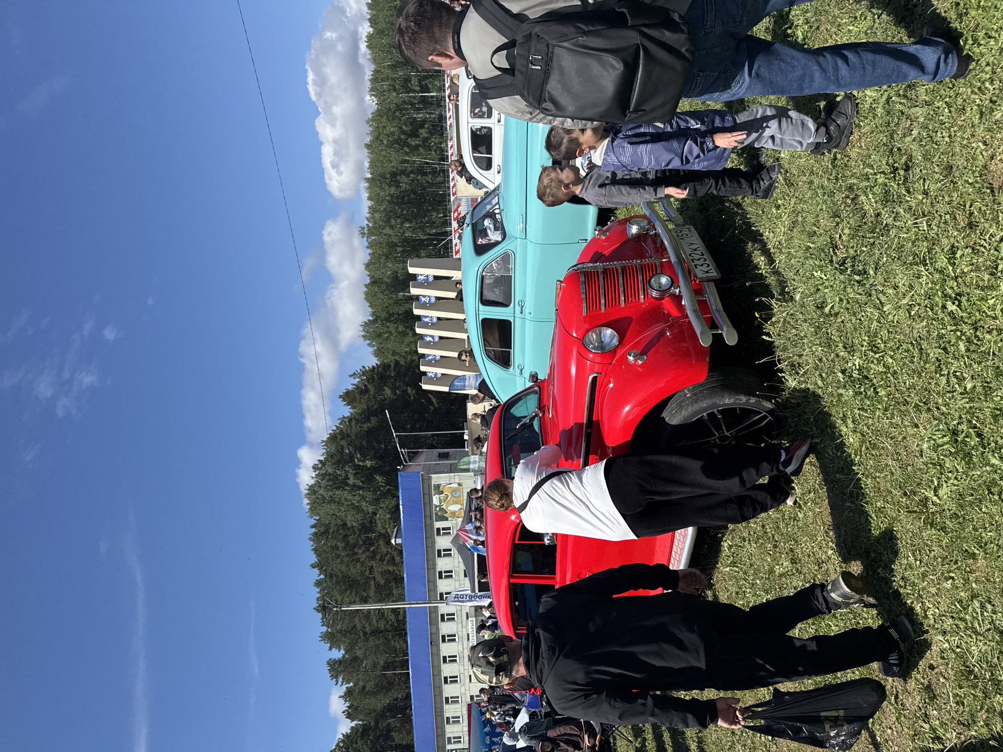 The image captures a scene at what appears to be a car show or exhibition in the city of Chaikovsky, Russia. Multiple vintage cars are lined up, including a red one and a blue one, with attendees inspecting them. The attendees include adults and children. Some attendees are looking at or touching the cars. A building is visible in the background, and the environment suggests a grassy outdoor area, possibly a park or fairground. The sky is blue with some clouds. The image is taken during daylight hours. Based on the license plate visible on the red car, the region is likely Perm Krai.