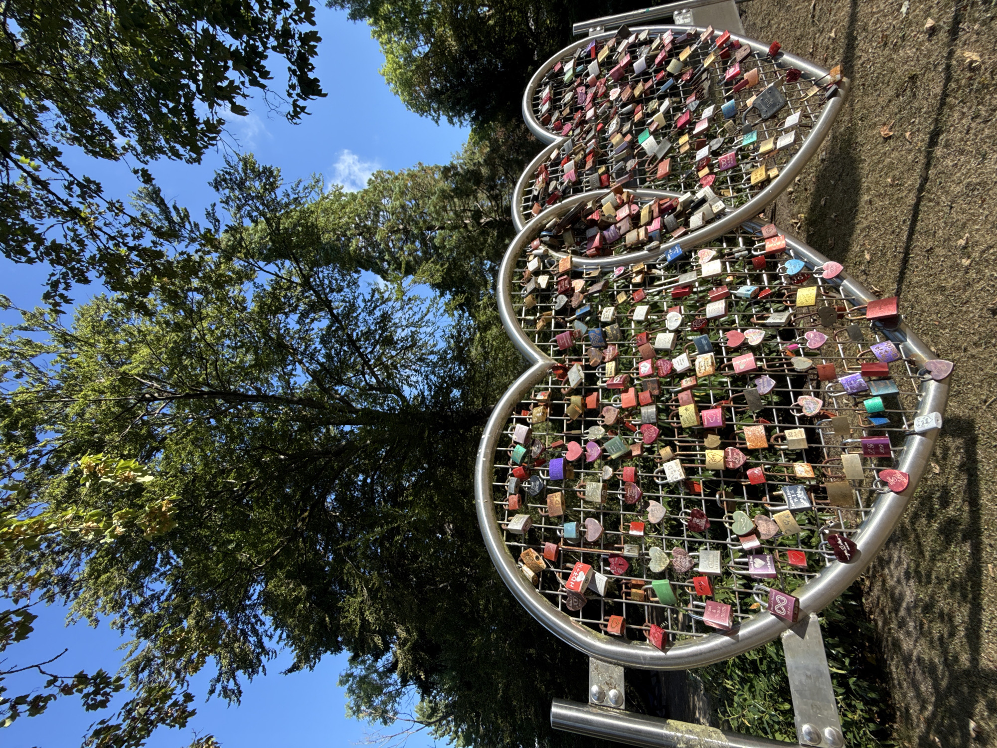 This image shows a sculpture made of two interconnected heart shapes, adorned with numerous padlocks. The sculpture is located in the town of Bad Sachsa, Germany. The padlocks are of various colors, shapes, and sizes, with some appearing to have engravings or writing on them. The sculpture is made of metal mesh and supported by metal posts, positioned outdoors in a park-like setting with trees and a grassy ground. The sky is blue with a few scattered clouds, and the sun is shining, casting shadows on the ground. No people are visible in the image.