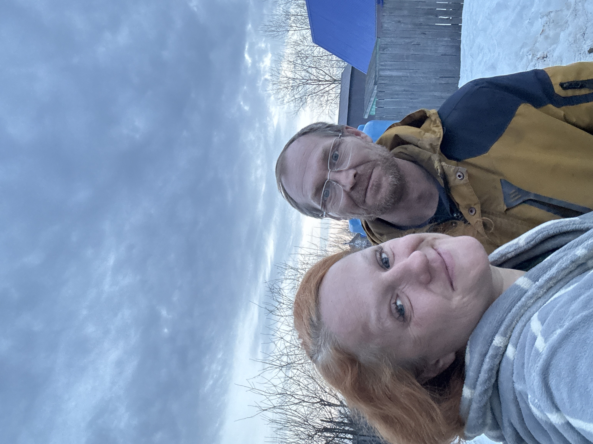 A photograph depicts two adult individuals outdoors in the city of Chaikovsky, Russia. In the foreground, positioned slightly to the left, is a woman with reddish-blonde hair, wearing a grey patterned fleece garment, smiling and looking directly at the camera. Behind her and slightly to the right stands a man with short brown hair, a beard, and eyeglasses, wearing a mustard yellow and dark blue winter jacket, also smiling and looking at the camera. The ground in the mid-ground and background is covered with snow, with some patches of visible earth. Behind the individuals, a wooden fence spans horizontally. Beyond the fence, several bare deciduous trees or bushes are visible. Further in the background, residential structures include a building with a dark roof and another with a prominent bright blue corrugated metal roof. The sky is overcast with grey and light blue clouds, indicating an outdoor setting with diffused lighting, suggestive of late winter or early spring conditions.