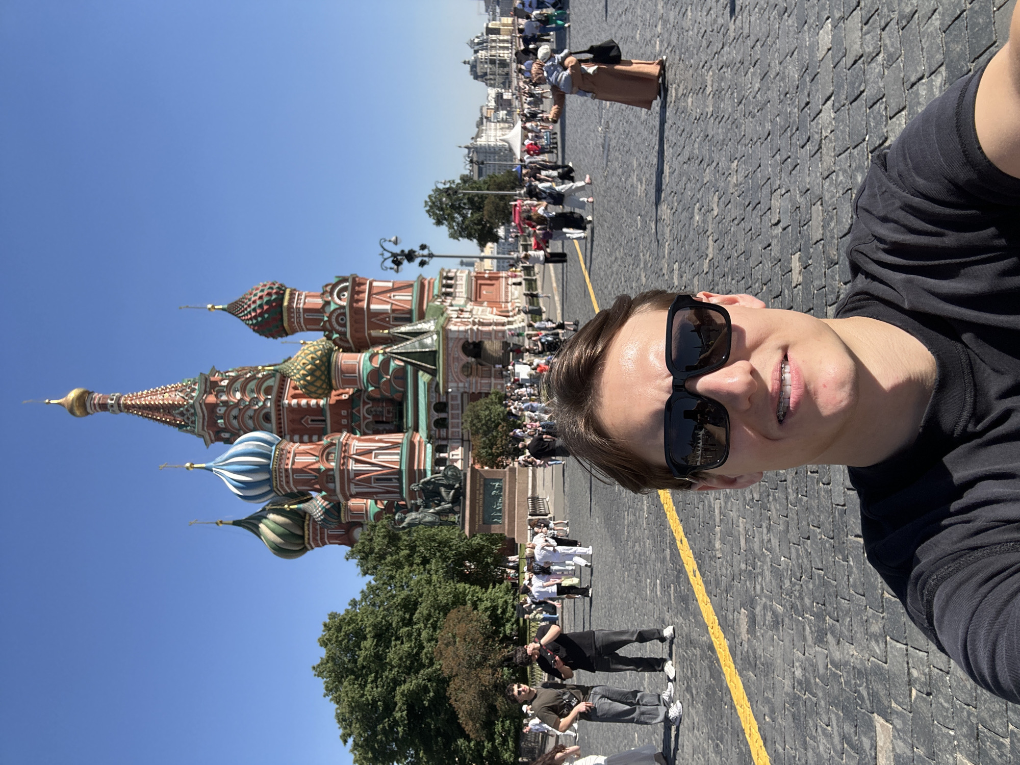 A young adult male is captured in a selfie at Red Square in Moscow, Russia, with St. Basil's Cathedral prominently featured in the background. The man, positioned in the lower-center foreground, has light skin, short brown hair, and is wearing dark rectangular sunglasses and a black crew-neck t-shirt. Braces are visible on his upper teeth as he smiles. The foreground consists of dark gray cobblestone pavement with a distinct yellow line running horizontally across the lower frame, disappearing behind the man.
Behind the man, St. Basil's Cathedral stands under a clear, bright blue sky. Its iconic multi-colored onion domes, featuring patterns of gold, blue and white stripes, green and white stripes, and red and green diamond patterns, are clearly visible atop the red brick structure. To the left of the cathedral, the Minin and Pozharsky monument is identifiable on its pedestal.
Numerous individuals are dispersed across Red Square, walking in various directions. On the left side of the frame, two men stand on the cobblestones; one wears a dark t-shirt and jeans, the other a khaki t-shirt and jeans, with a camera strap visible around his neck. On the right, a person in a long, light brown robe holds a small child. Green trees are visible in the midground to the left, behind groups of people. Buildings of the distant Moscow skyline are faintly discernible on the far right horizon. The scene is illuminated by bright daylight, casting visible shadows.