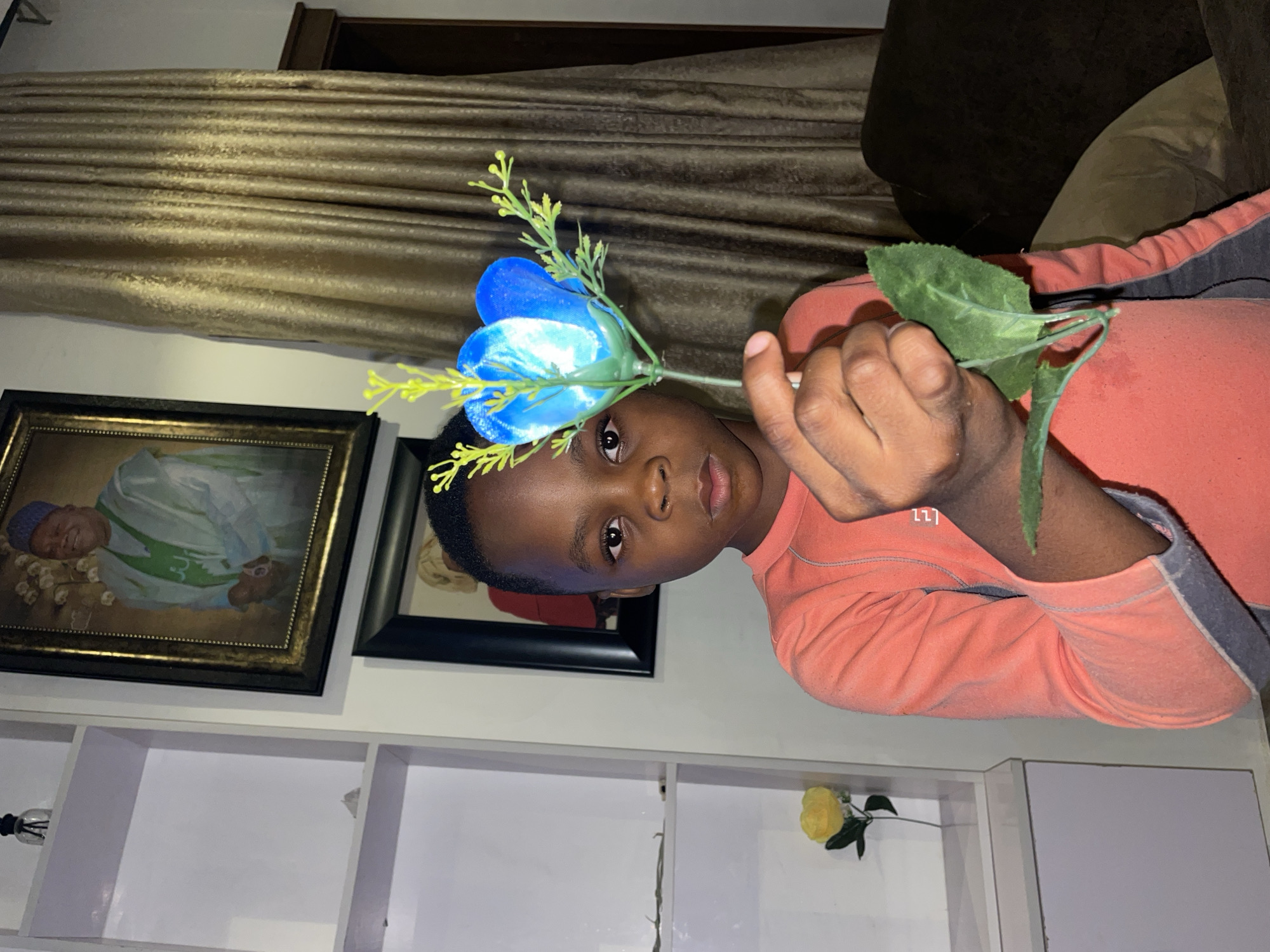 A young boy in an orange shirt holds a blue artificial flower. He is inside a room with white shelves, on which are a diffuser and a yellow artificial flower. On the wall behind him are two framed pictures and a gold-colored curtain. The location is Jos, Nigeria.