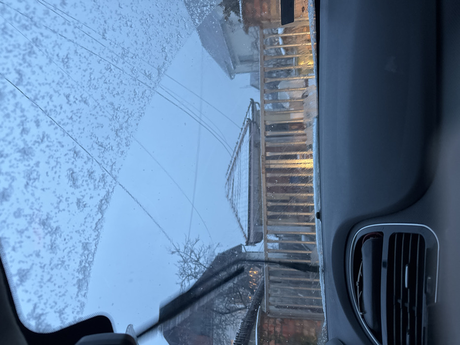 The image depicts a winter scene in Cibukovac, Serbia, observed from the interior of a vehicle through its front windshield. The windshield is partially obscured by frozen precipitation (snow/ice) across the upper section and contains visible cracks. A dark-colored windshield wiper blade is visible on the left, appearing in motion or recently activated. The vehicle's dark gray dashboard, featuring an air vent on the left, occupies the lower foreground.
Beyond the windshield, a light-colored wooden fence or railing extends horizontally across the mid-ground. Behind the fence, several buildings are visible. To the left, a portion of a residential building with a dark roof and a bare, leafless tree stands. Centrally, a smaller structure with a snow-dusted, reddish-brown tiled roof is present. Another residential building with a snow-covered roof is partially visible on the right. Overhead, multiple utility lines crisscross the overcast, light blue-grey sky. A localized warm, yellow light source is visible behind the fence in the mid-right, casting reflections on the fence slats and ground. No people are visible.