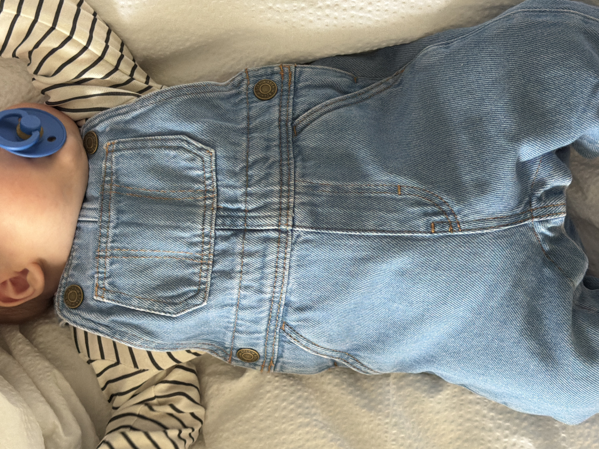 A baby is captured in a close-up, high-angle shot, lying on a white, textured surface, likely a blanket or sheet. Only the left side of the baby's head and ear are visible, with light skin tone. A bright blue, round-mouthed pacifier is in the baby's mouth.
The baby is dressed in a long-sleeved shirt with horizontal black and off-white stripes, over which light blue denim overalls are worn. The denim features contrasting light brown or orange stitching, visible on seams and pockets. Antique bronze-colored buttons adorn the overalls; one prominently displays the engraved text "AUTHENTIC DENIM." The baby's body is mostly covered by the overalls, which appear soft and slightly creased, indicating comfort. The overall impression is one of a peaceful, resting baby in an indoor setting.