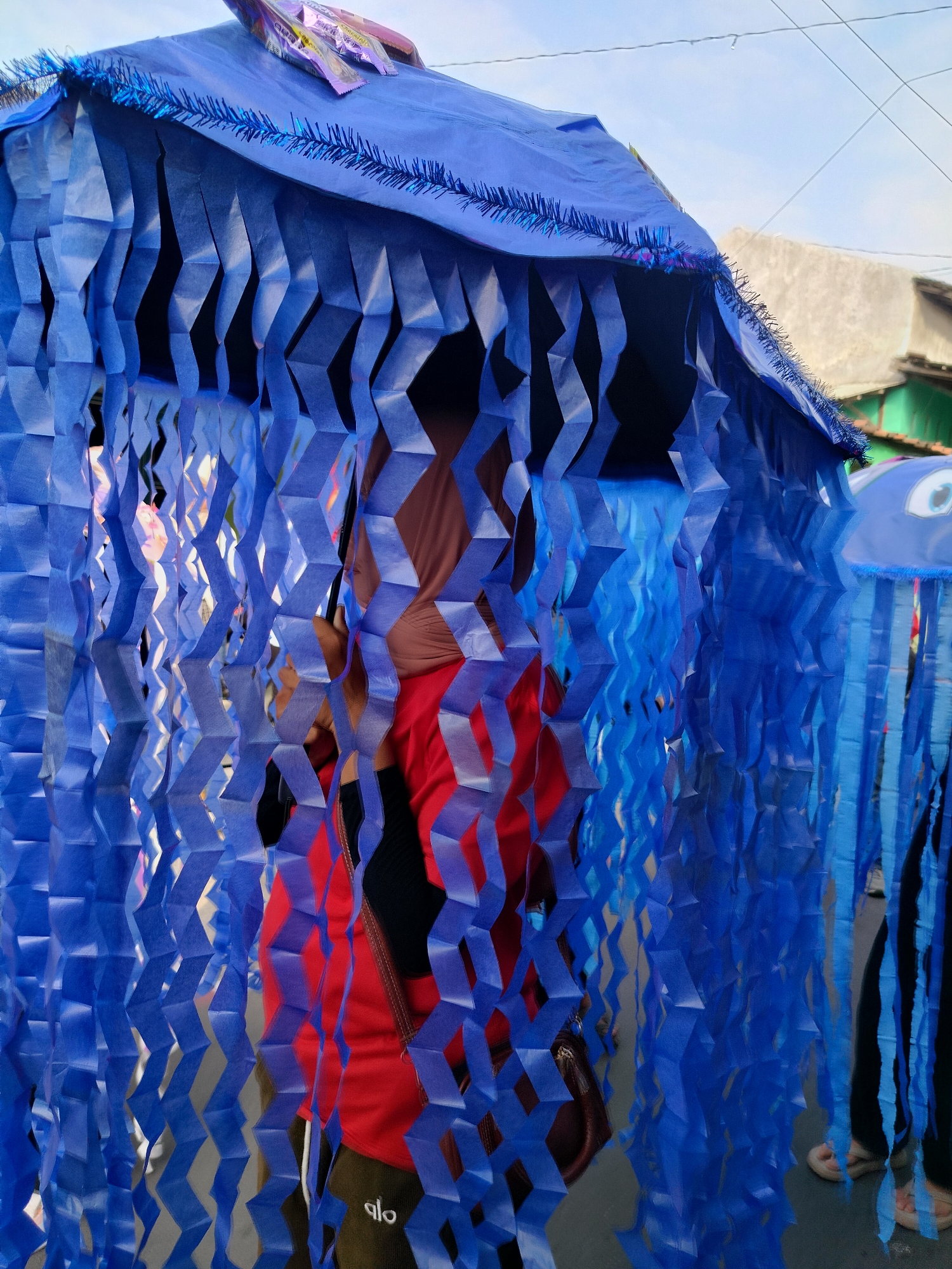 The image shows a close-up view of a blue, tent-like structure adorned with hanging, blue paper strips, likely part of a festival or parade float. The structure is decorated with a blue tinsel trim along its roofline and appears to have a roof made of blue plastic sheeting. Behind the blue paper fringe, a person wearing a brown hijab and a red shirt is visible, along with a dark bag slung over their shoulder. The ground is paved, and in the background, a building with a white wall and a green building are discernible, suggesting an outdoor setting in a town. A similar blue decoration, possibly another float or part of the same display, with a large eye visible, is to the right of the frame. The scene is likely from a street in Juwiring, Indonesia, given the context.