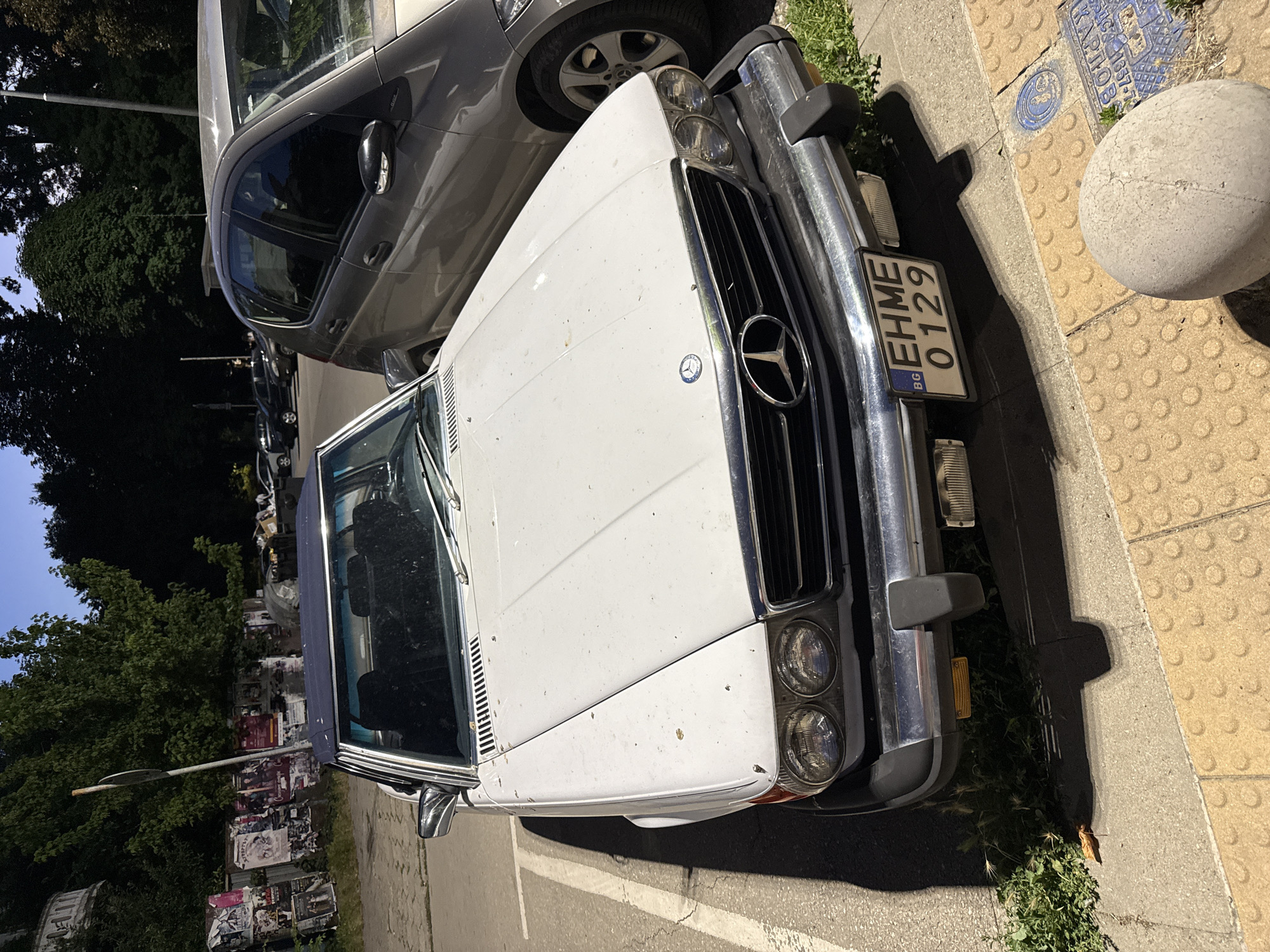 A white Mercedes-Benz SL-Class convertible is parked on a street in Pleven, Bulgaria. The car has a dark blue convertible top and a Bulgarian license plate that reads "BG EHME 0129". The car is parked next to a grey hatchback and a sidewalk with tactile paving. In the background, there are trees, posters on a fence, and other parked cars. The scene appears to be in an urban area.