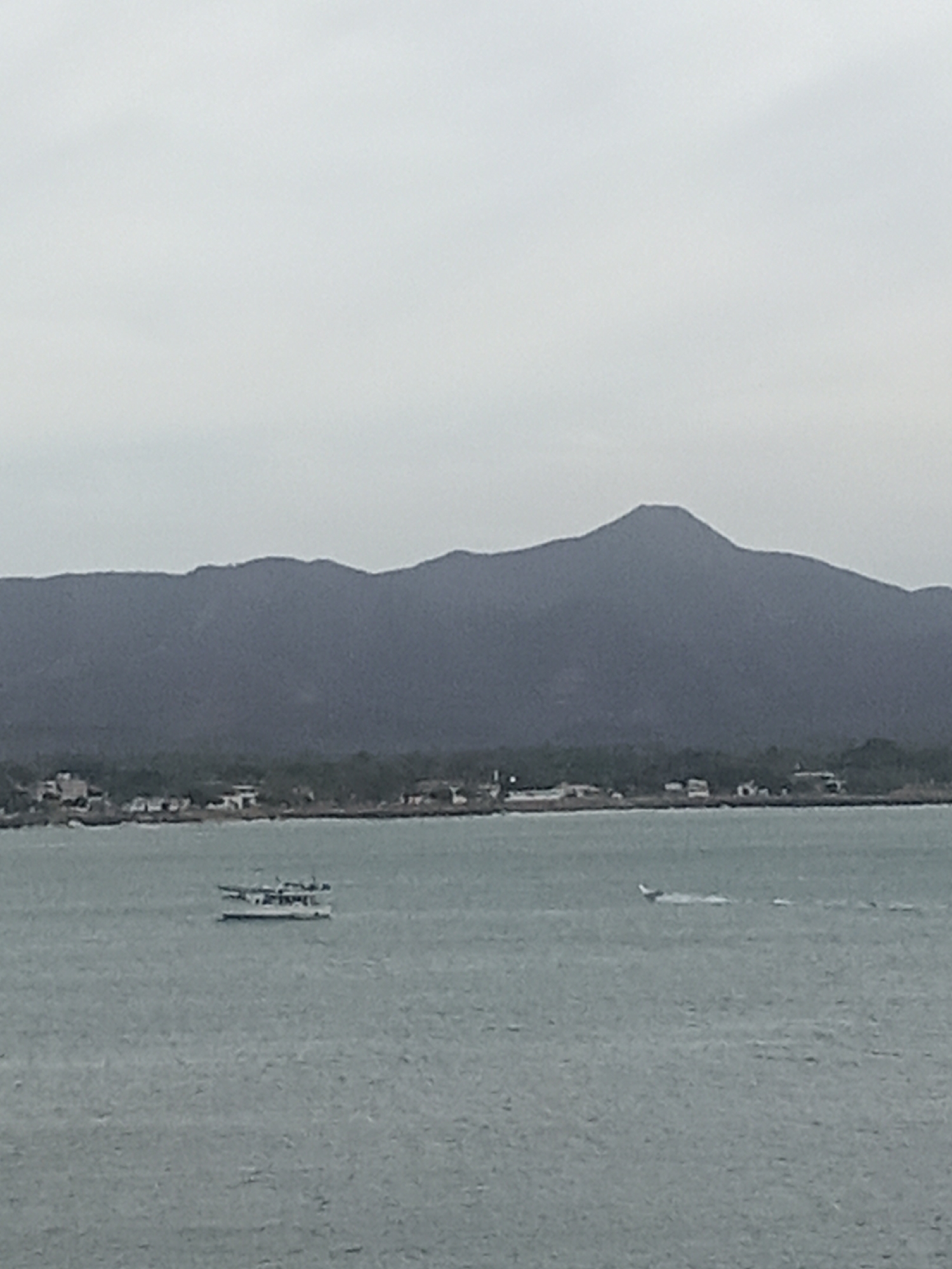 The image depicts a seascape under an overcast sky. In the foreground, two vessels are visible on the water. One appears to be a small boat with passengers, while the other is a seaplane or similar aircraft skimming the surface. In the background, a coastline is discernible with buildings and foliage along the shore. Further back, a mountainous terrain rises. Given the location, this may be a view in the city of Juan Griego, Venezuela.
