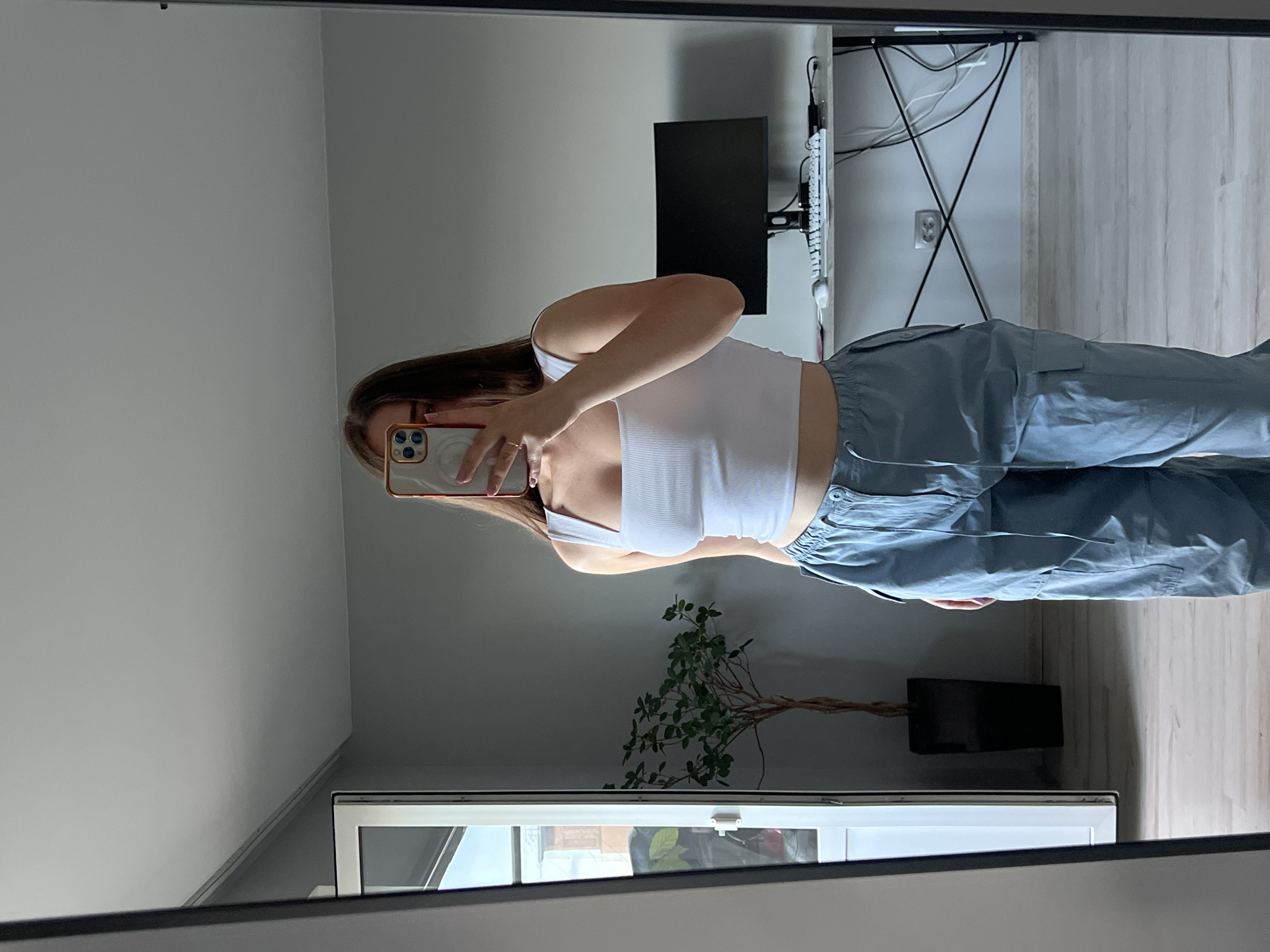 A young woman is taking a mirror selfie in a room. She is wearing a white ribbed tank top and light blue cargo pants. She is holding a phone in her right hand, and her face is obscured by the phone. In the background, there is a large mirror, a potted plant, a desk with a computer monitor and keyboard, and a doorway leading to another room or outdoor area. The room appears to be a bedroom or living space with light gray walls and light wood flooring. The location of the image is in the city of Ternopil, Ukraine.