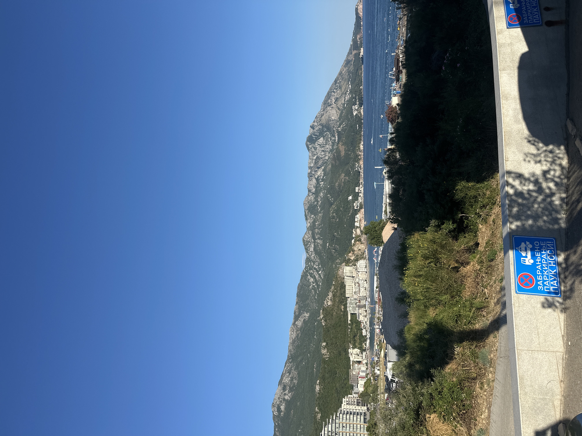This is a medium-shot, eye-level, outdoor, daytime photograph of the coastal city of Boreti, Montenegro, taken on a clear day. The image captures a scenic view of the Adriatic Sea, with mountains in the background and a portion of the city in the midground. The foreground is dominated by a low, light-colored stone wall with a blue and white traffic sign on it. The sign features a tow truck icon and text in Cyrillic, indicating "No Parking, Tow Away Zone." Shadows of trees and possibly people are cast on the wall and the paved surface in front of it.
Behind the wall, lush green vegetation and trees are visible, with some dry, yellowed grass and bushes. Further back, a densely built urban area lines the coast, featuring numerous white and light-colored buildings, including hotels and apartment complexes, nestled against the verdant foothills of the mountains. The mountains themselves are covered in dense green foliage, with rocky outcrops and peaks visible towards the top. The calm, blue waters of the Adriatic Sea stretch out towards the horizon, with several boats and some small structures visible along the coastline. The sky is a clear, bright blue. The overall impression is one of a sunny day in a popular tourist destination.
