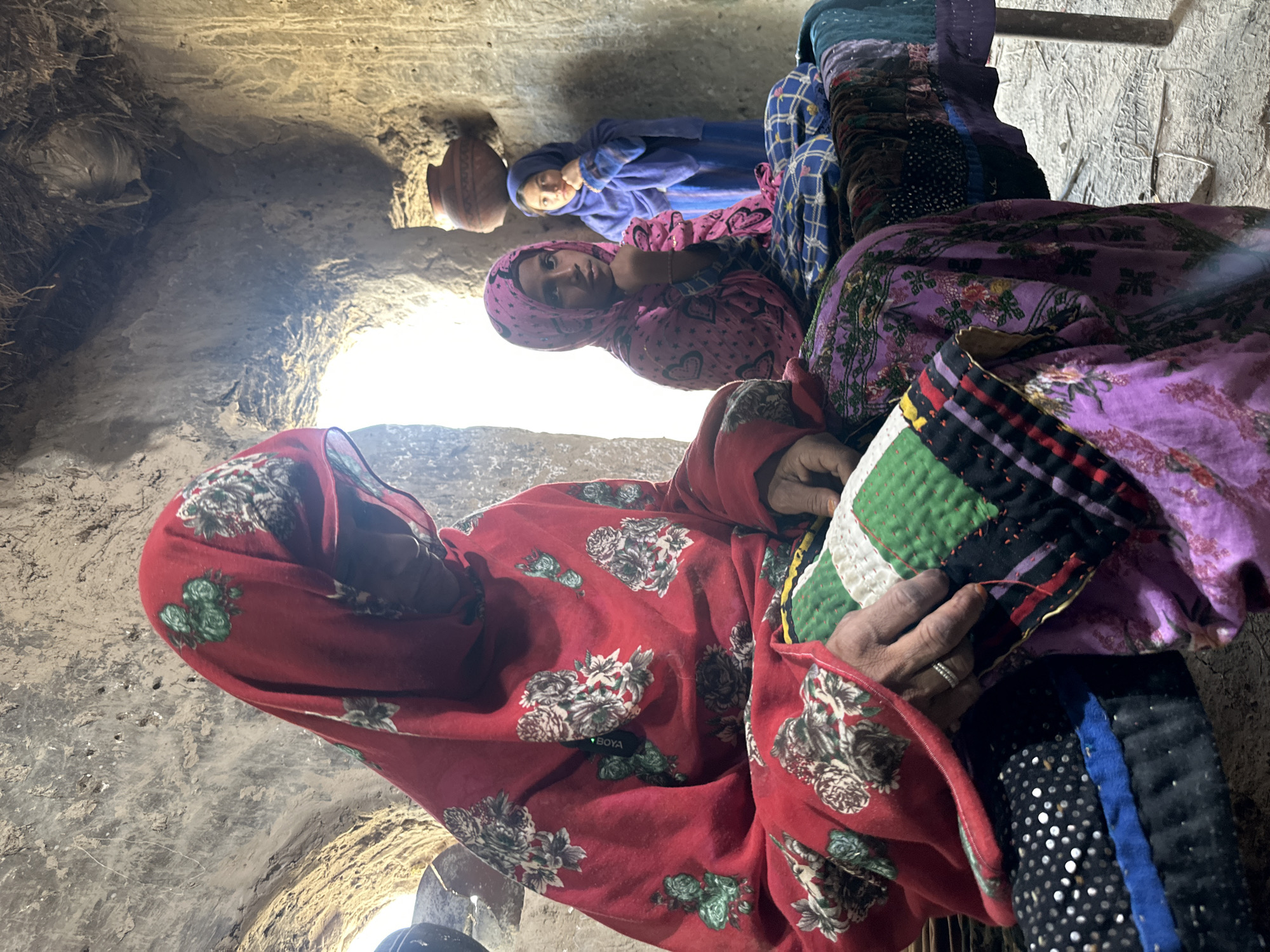 The image, likely taken in Quetta City Tehsil, Pakistan, features three women indoors, possibly in a dwelling with a rough, earth-like interior. The woman in the foreground is wearing a red patterned dress with a matching headscarf. She is seated and appears to be sewing a patchwork quilt. Behind her, a woman in a pink patterned outfit and headscarf looks toward the camera. In the background, another woman in a blue patterned dress and headscarf is seated. A clay pot sits on a shelf or ledge in the background. The environment appears modest and somewhat dimly lit, with a light source visible in the top left corner. The overall impression is one of domesticity and traditional activity.