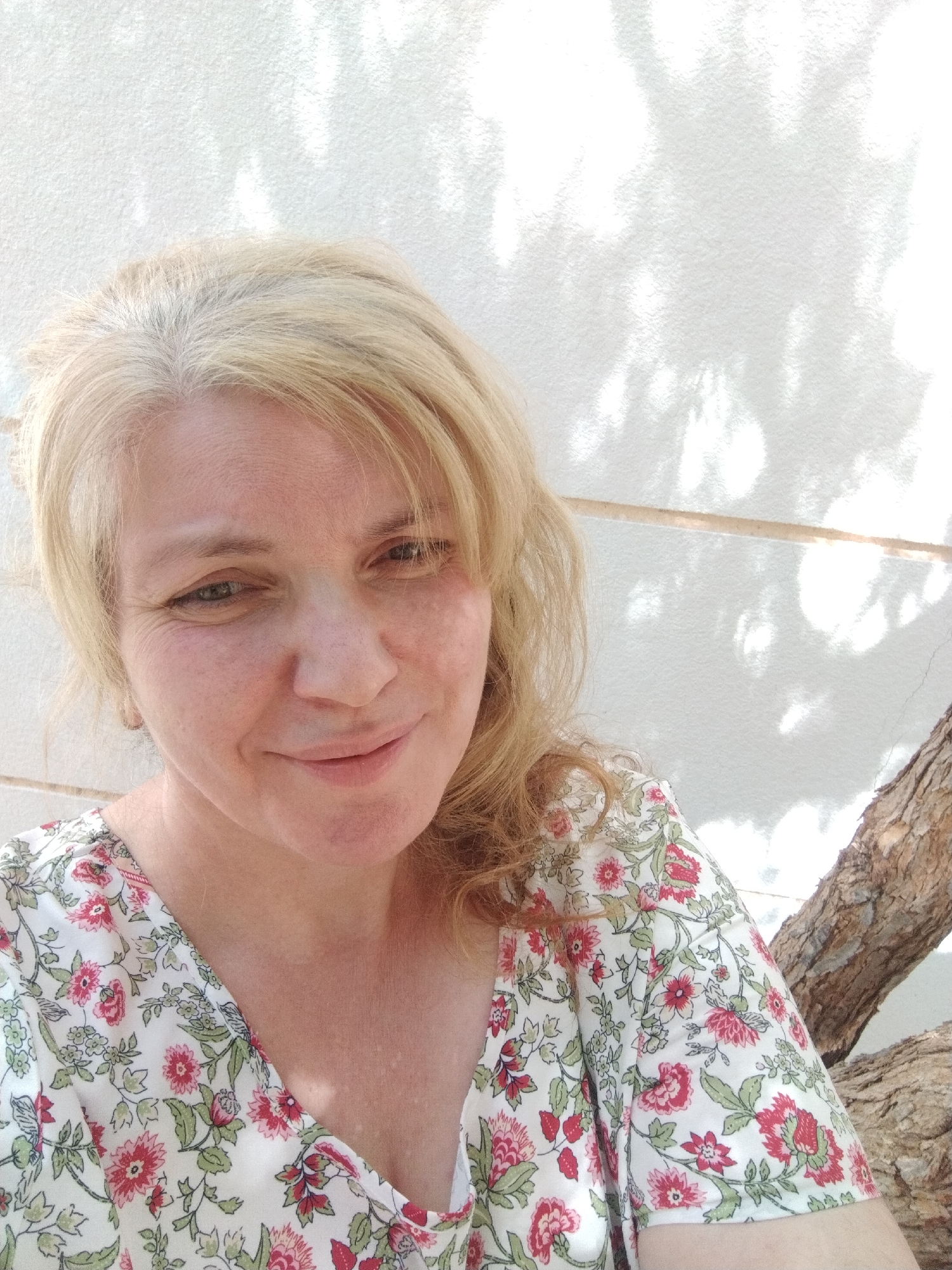 The photograph captures a fair-skinned woman with blonde hair, exhibiting gray roots. She wears a floral-patterned top, featuring red and green flowers on a white background. The backdrop is a white textured wall, partially obscured by the shadow of leaves. A tree branch extends into the frame from the right side. The photograph was taken in Al Mas'oudi, United Arab Emirates.