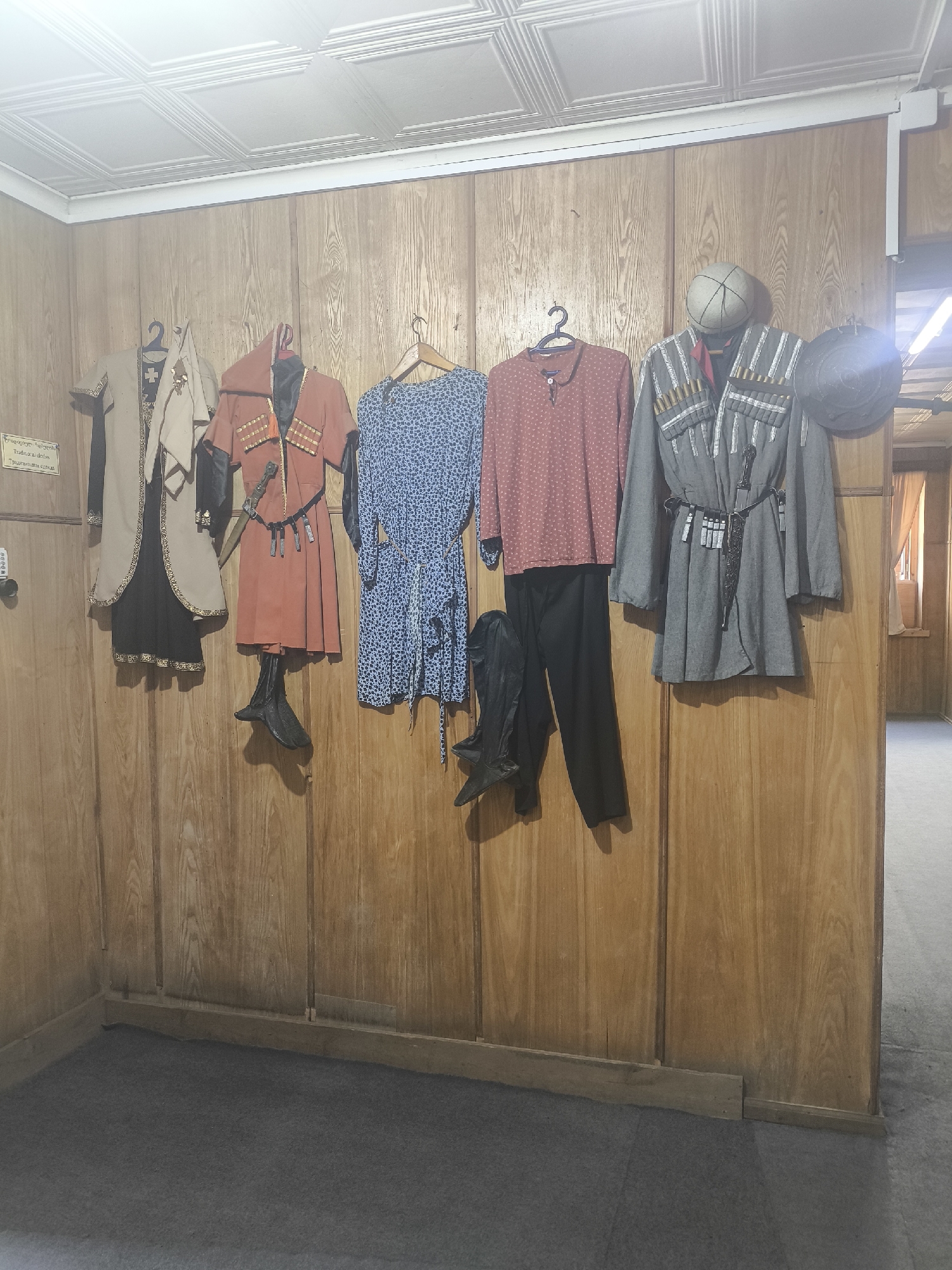 The image displays a collection of traditional Georgian attire displayed on mannequins against a wooden paneled wall. The attire includes a long beige coat with black trim over a black skirt, a red tunic with ornate trim and a dagger tucked into a belt, a blue patterned dress, a red patterned long-sleeved shirt paired with black pants, and a grey military-style jacket adorned with bullet cartridges and a dagger. Boots are also displayed. A sign in Georgian script is visible on the wall to the left. The scene appears to be an exhibit or display in a museum or cultural center, likely located in Batumi, Georgia, given the presence of traditional Georgian clothing. The lighting suggests an indoor setting.