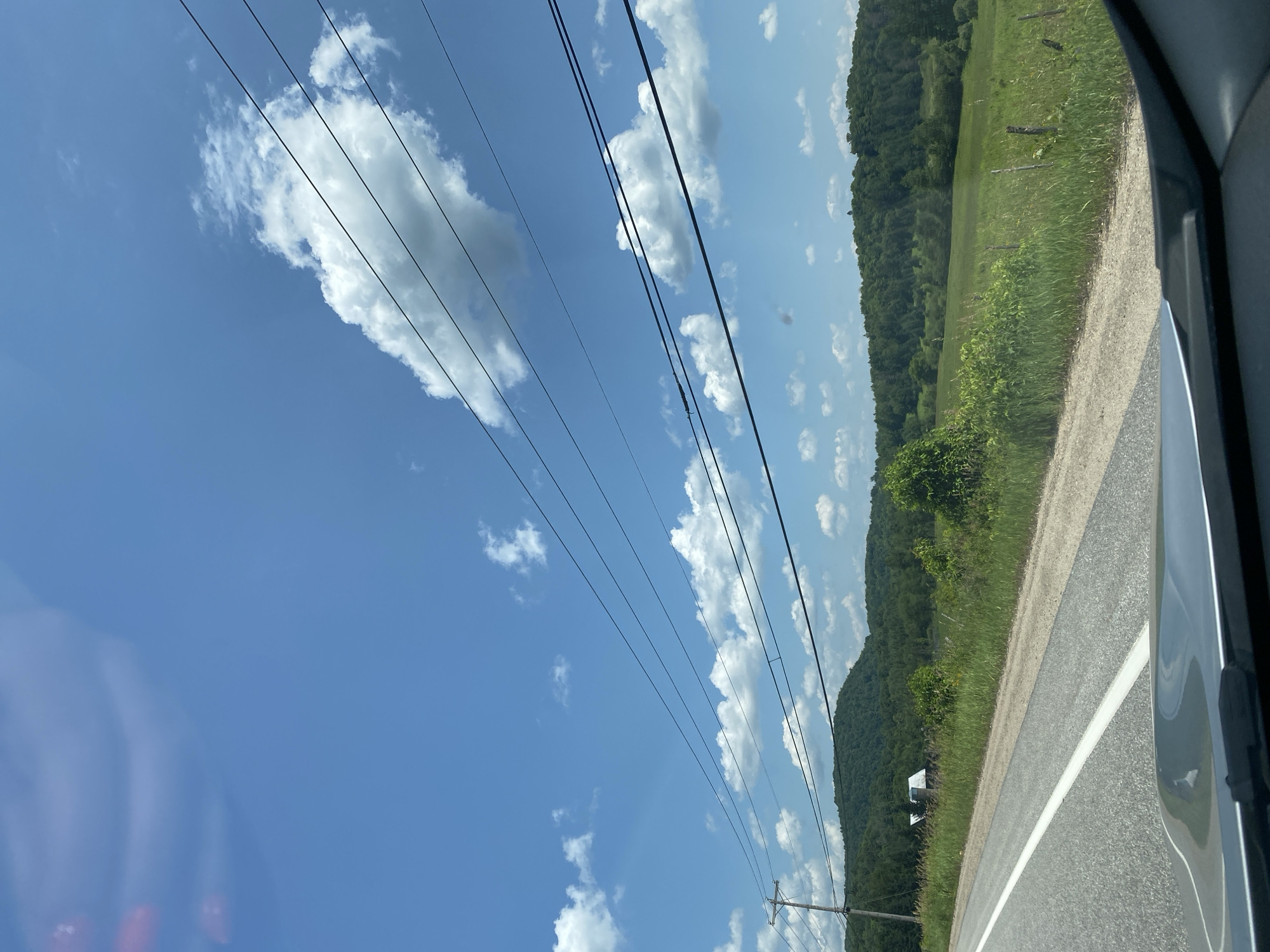 The image depicts a rural roadside scene under a bright blue sky with scattered white clouds. A paved road with a white line runs diagonally from the lower left to the middle right of the frame. To the right of the road is a grassy field with scattered wildflowers and a few wooden fence posts. Beyond the field, a treeline of green deciduous trees forms a gentle slope towards a distant, larger, forested hill. In the middle distance, nestled amongst the trees on the left side of the road, is a white building with a darker roof, possibly a barn or small house. Overhead, multiple power lines stretch across the sky, supported by a utility pole visible on the left side of the road. The perspective suggests the photo was taken from inside a vehicle, with the dashboard visible in the foreground. The environment is natural and undeveloped, characteristic of a countryside setting. There are no people or animals visible in the frame. The time of day appears to be daytime, with ample sunlight.